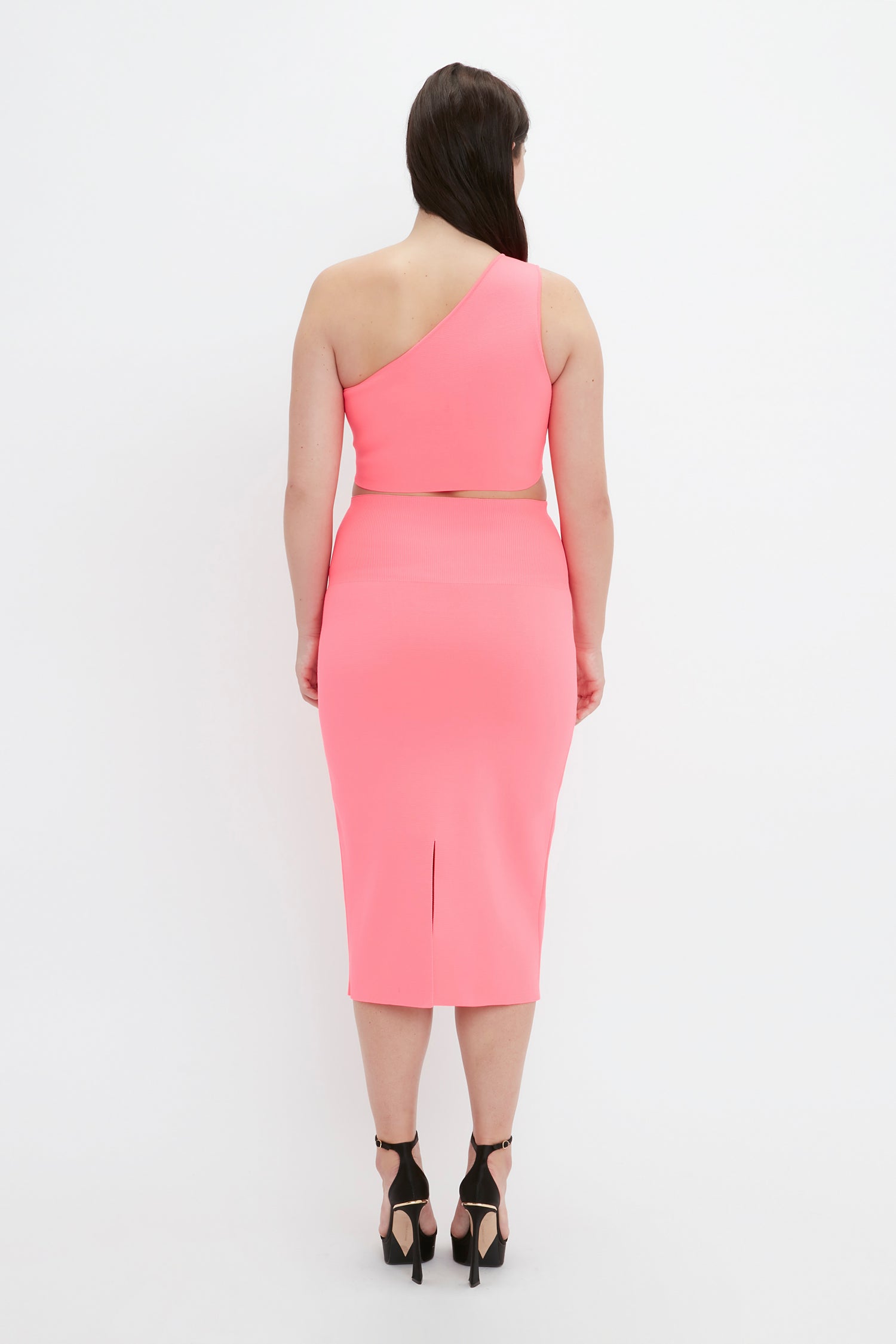 A woman is standing facing away, wearing the VB Body One Shoulder Crop Top in Pink by Victoria Beckham along with a matching pink skirt featuring a slit. She completes her outfit with black high-heeled sandals.