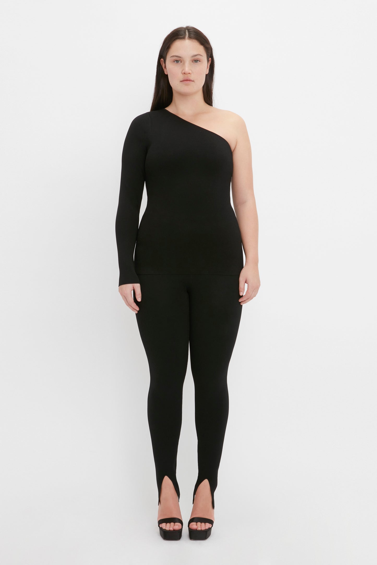 A person stands facing the camera, wearing the VB Body One Shoulder Top in Black by Victoria Beckham paired with black fitted pants, against a white background.