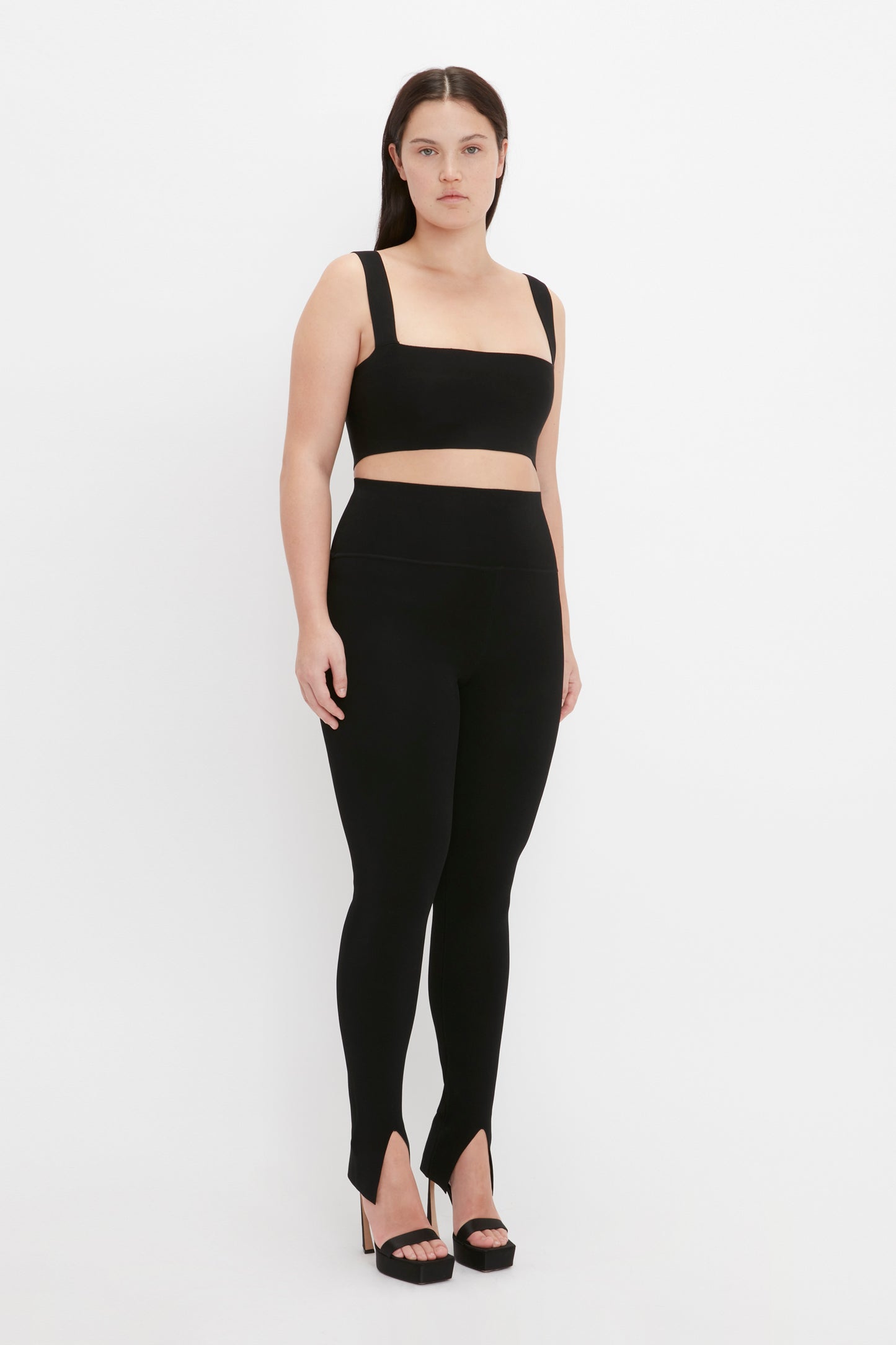 A woman stands against a plain white background, wearing a black sleeveless crop top, VB Body Split Front Leggings in Black by Victoria Beckham, and black high-heeled shoes.