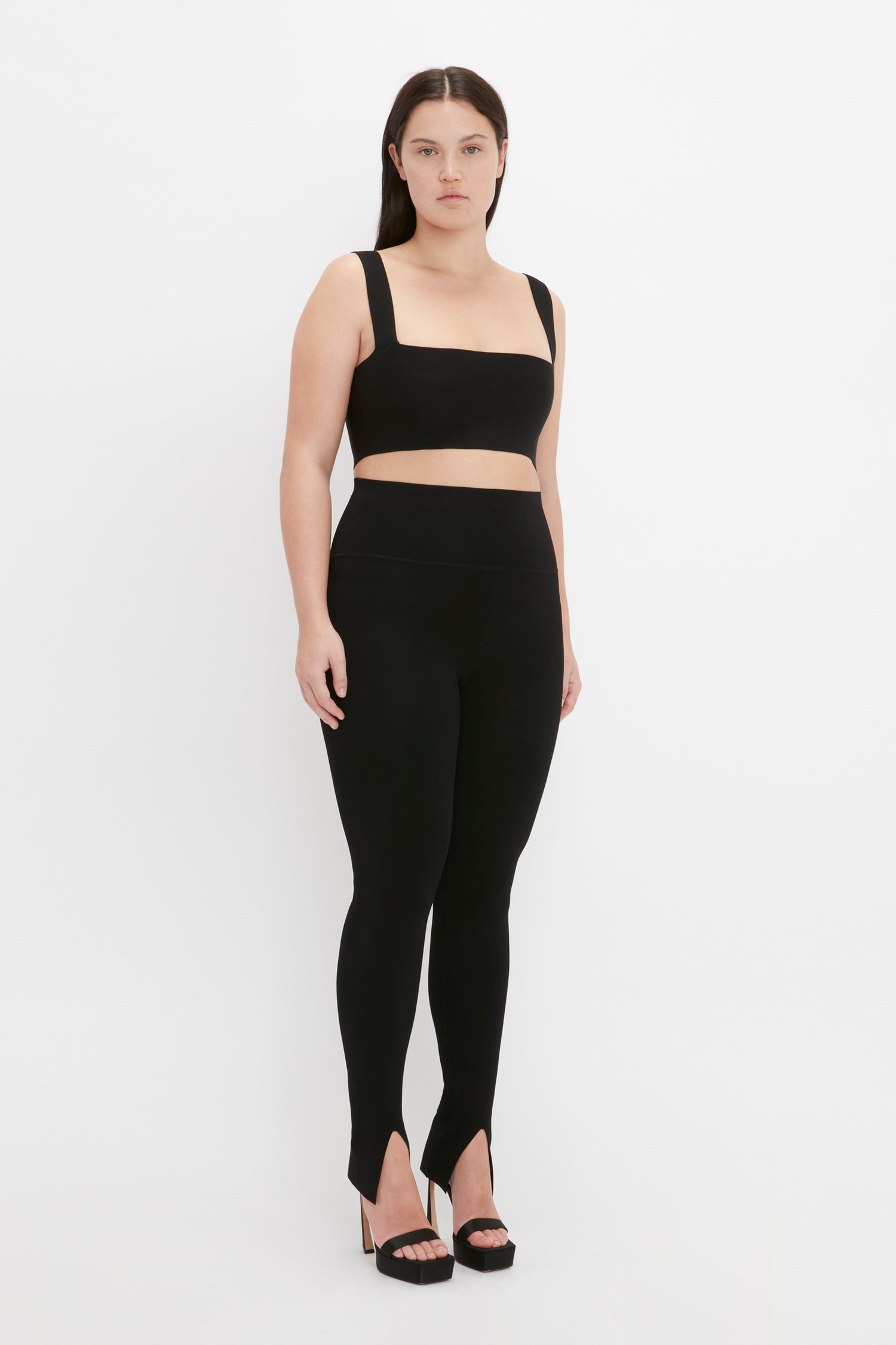 A woman stands against a plain white background, wearing a black sleeveless crop top, VB Body Split Front Leggings in Black by Victoria Beckham, and black high-heeled shoes.