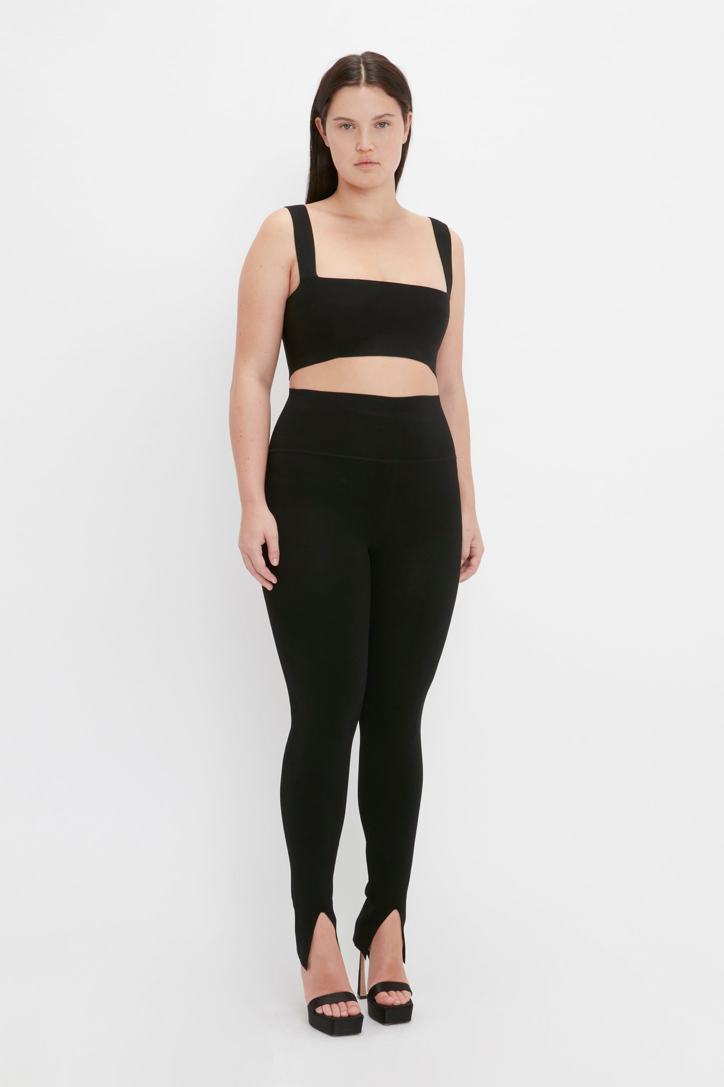 A person wearing the VB Body Strap Bandeau Top in black from Victoria Beckham, paired with high-waisted leggings and black high heels, stands against a plain white background.