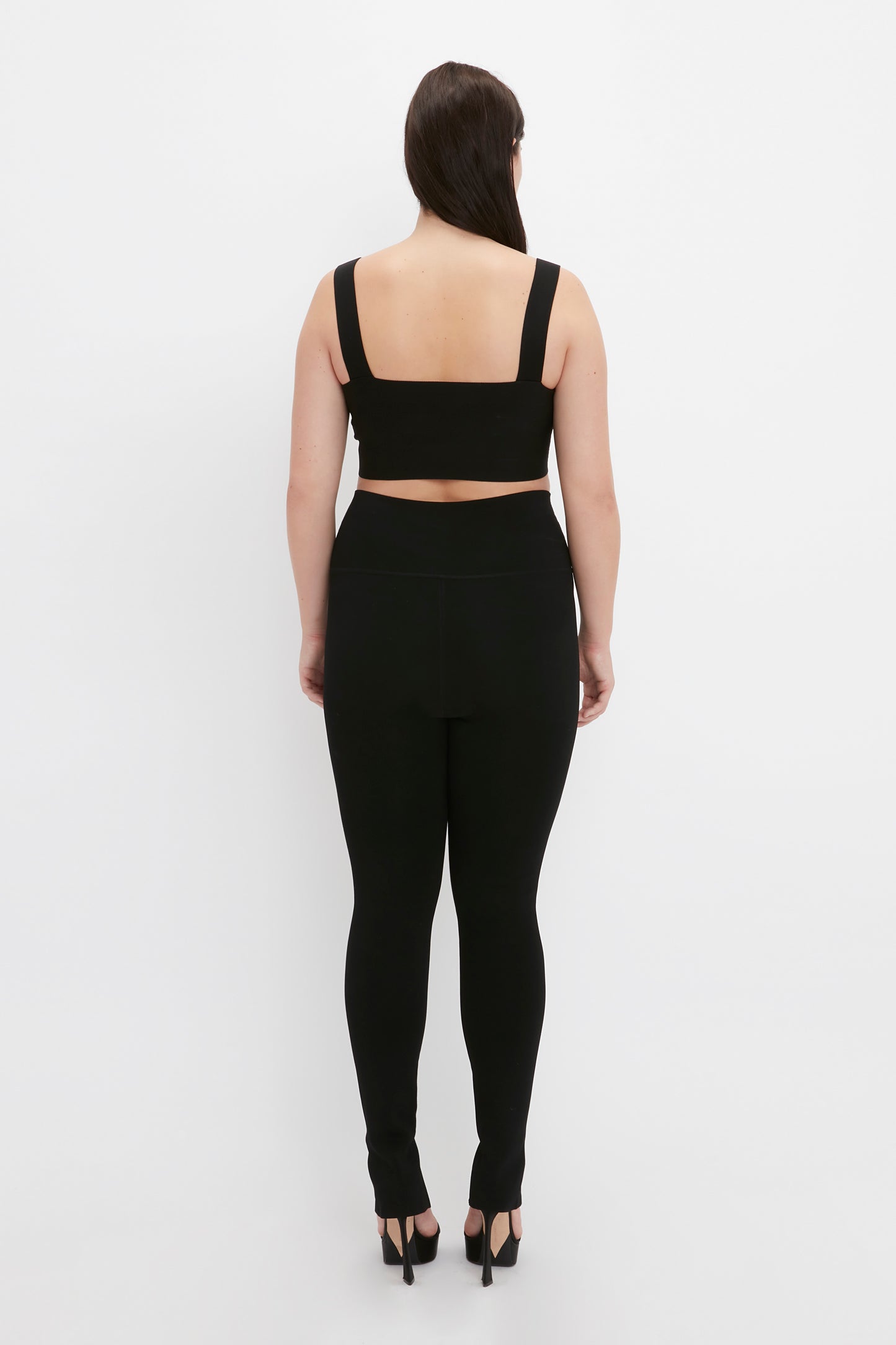 A person is standing facing away from the camera, wearing a black sleeveless top, Victoria Beckham VB Body Split Front Leggings in Black, and black high-heeled shoes.