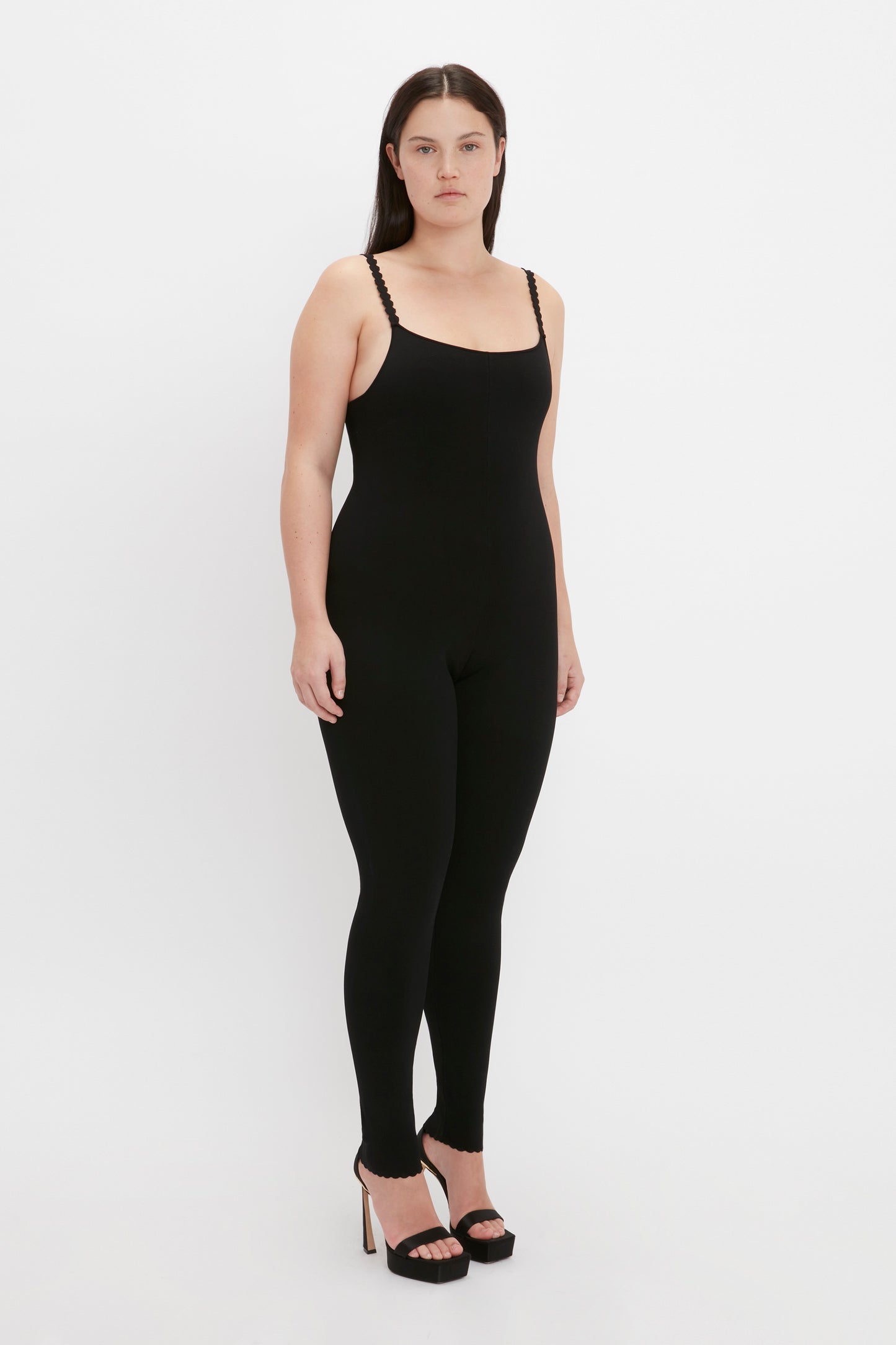 A person wearing the VB Body Catsuit in Black by Victoria Beckham, standing against a plain white background and wearing high-heeled shoes.