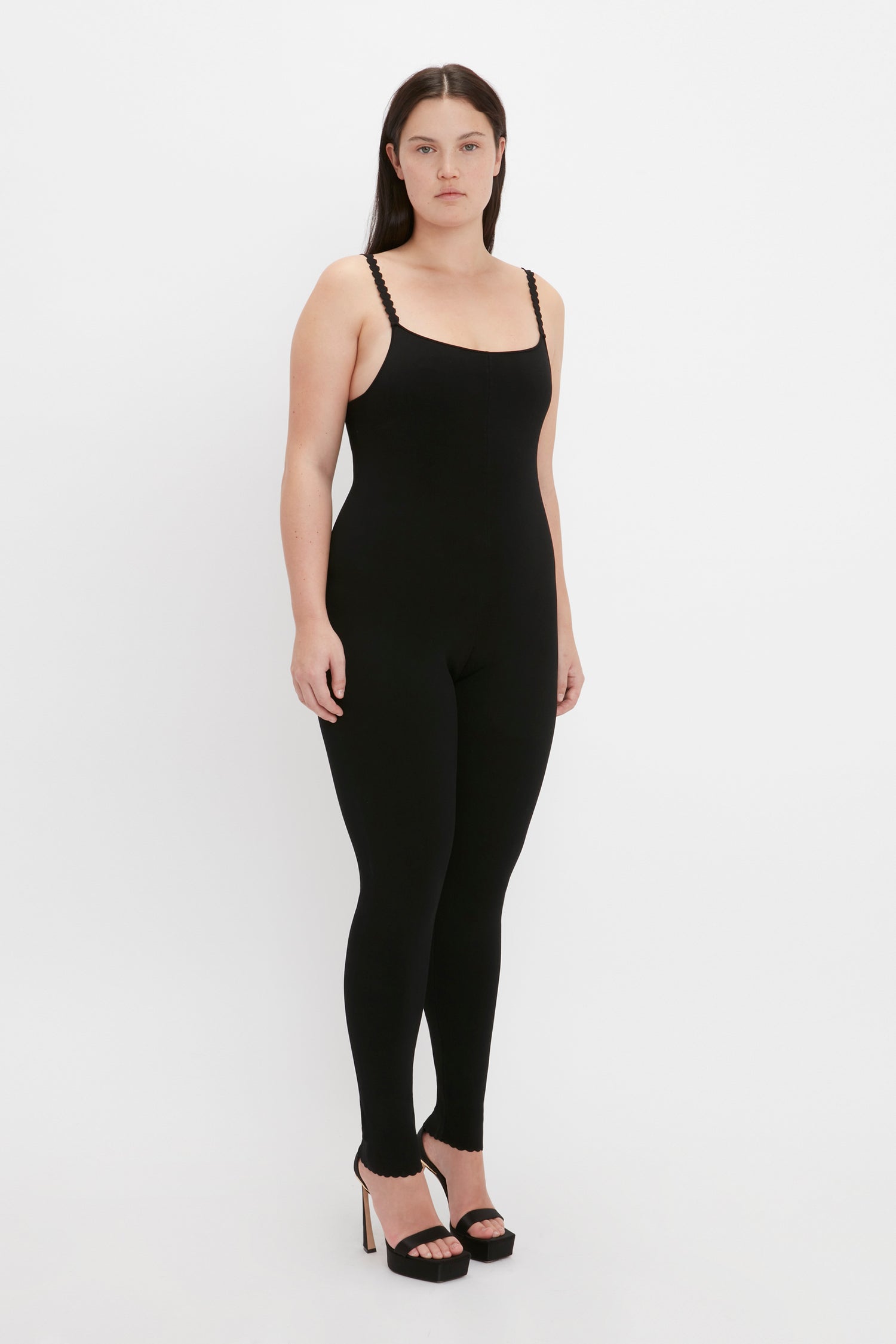 A person wearing the VB Body Catsuit in Black by Victoria Beckham, standing against a plain white background and wearing high-heeled shoes.