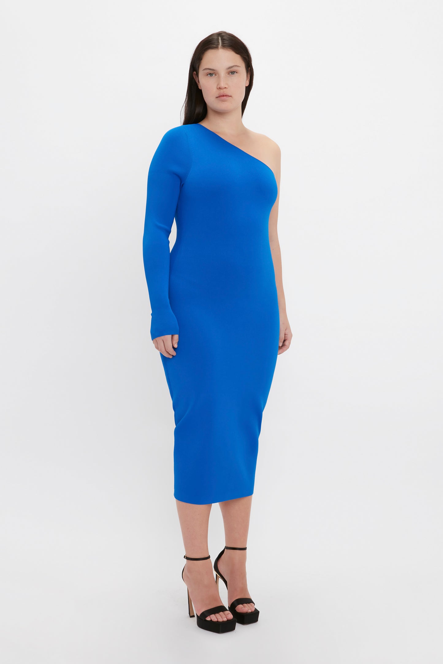 A person wearing the Victoria Beckham VB Body One Shoulder Midi Dress in Cobalt and black high heels stands against a plain white background.