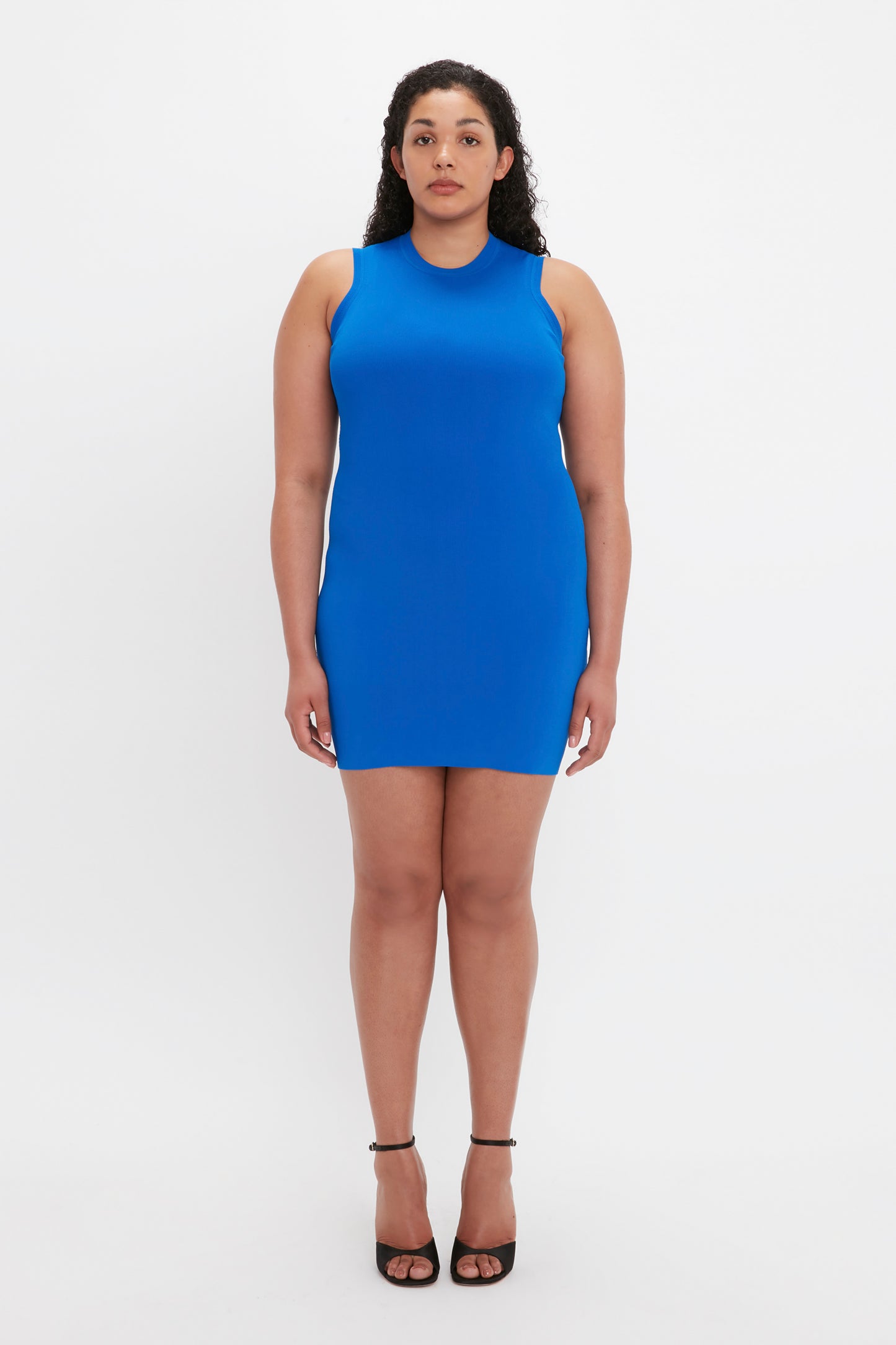 A person stands against a plain white background, wearing the VB Body Mini Dress in Cobalt by Victoria Beckham and black heels. Their arms are relaxed by their sides.
