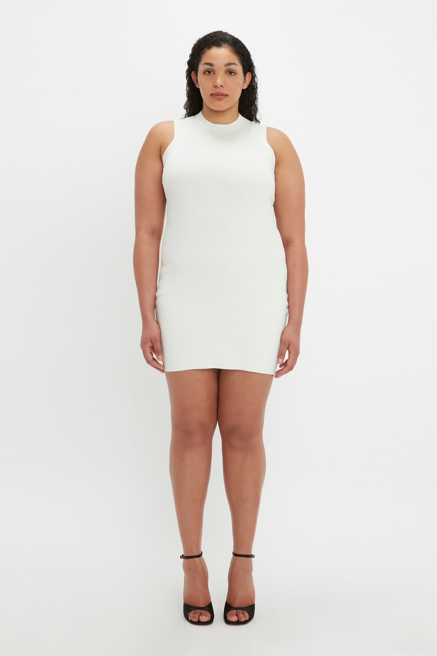 Person standing against a white background, wearing the VB Body Mini Dress in White by Victoria Beckham and black high-heeled shoes, looking directly at the camera with a neutral expression.