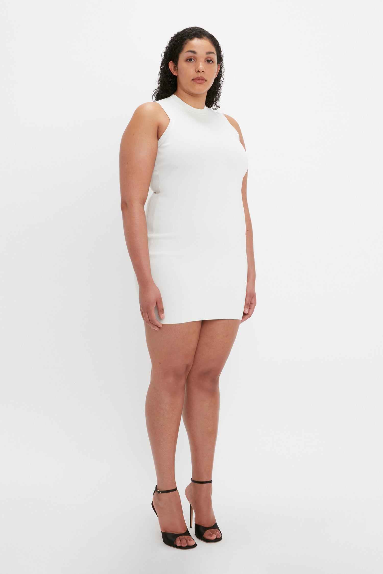 A person with curly hair stands against a plain white background wearing the VB Body Mini Dress in White by Victoria Beckham, paired with black high-heeled shoes.