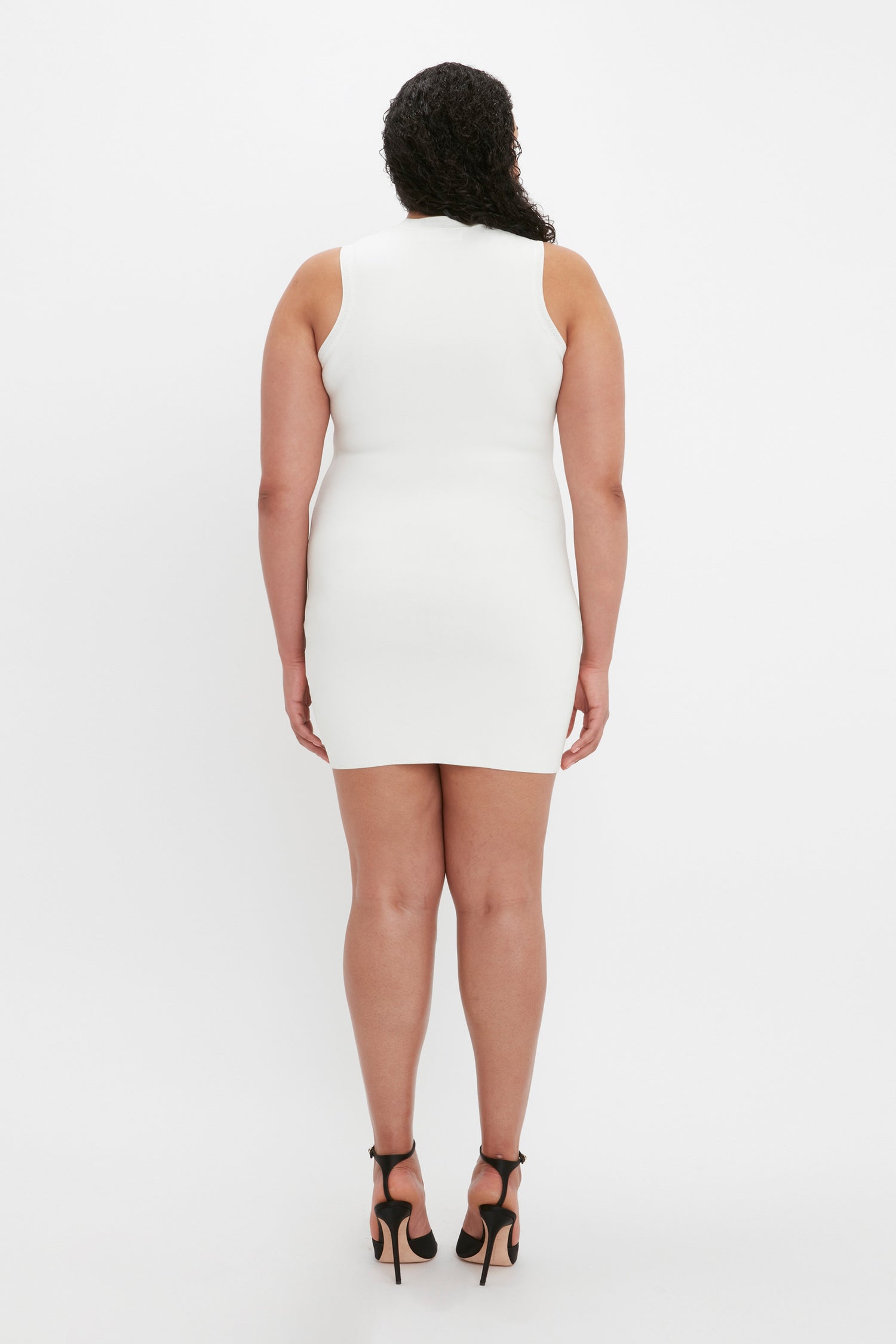 A person with curly hair stands facing away, wearing the VB Body Mini Dress in White from Victoria Beckham and black high heels against a plain white background.