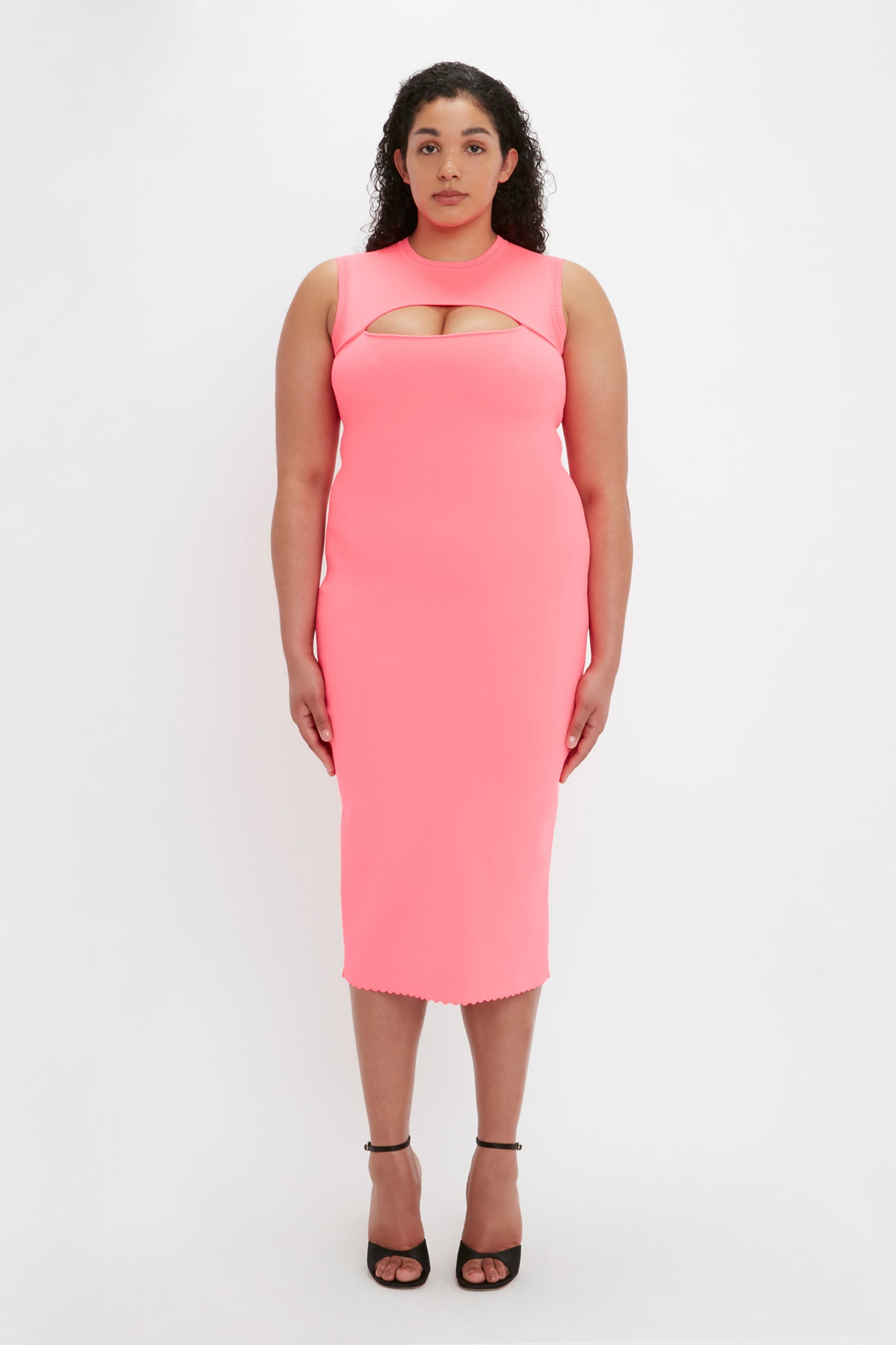 A woman with curly hair wears the Victoria Beckham VB Body Slash Front Midi Dress in pink paired with black high heels, standing against a white background.