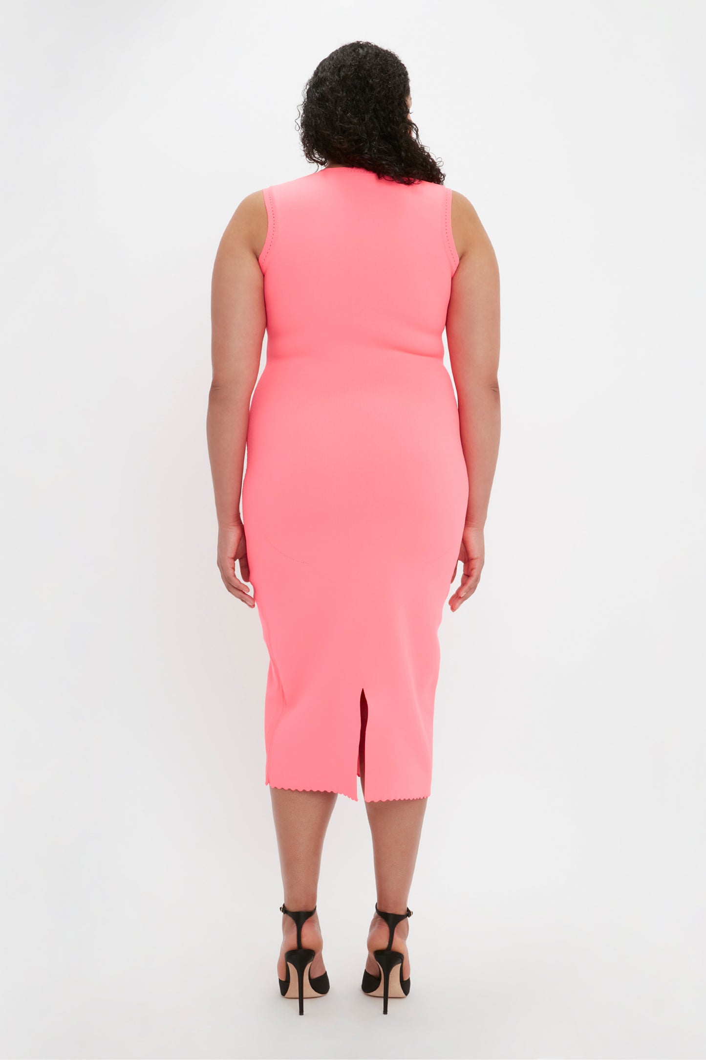 A person with curly hair is standing with their back to the camera, wearing a sleeveless, knee-length VB Body Slash Front Midi Dress in pink by Victoria Beckham and black high-heeled sandals.