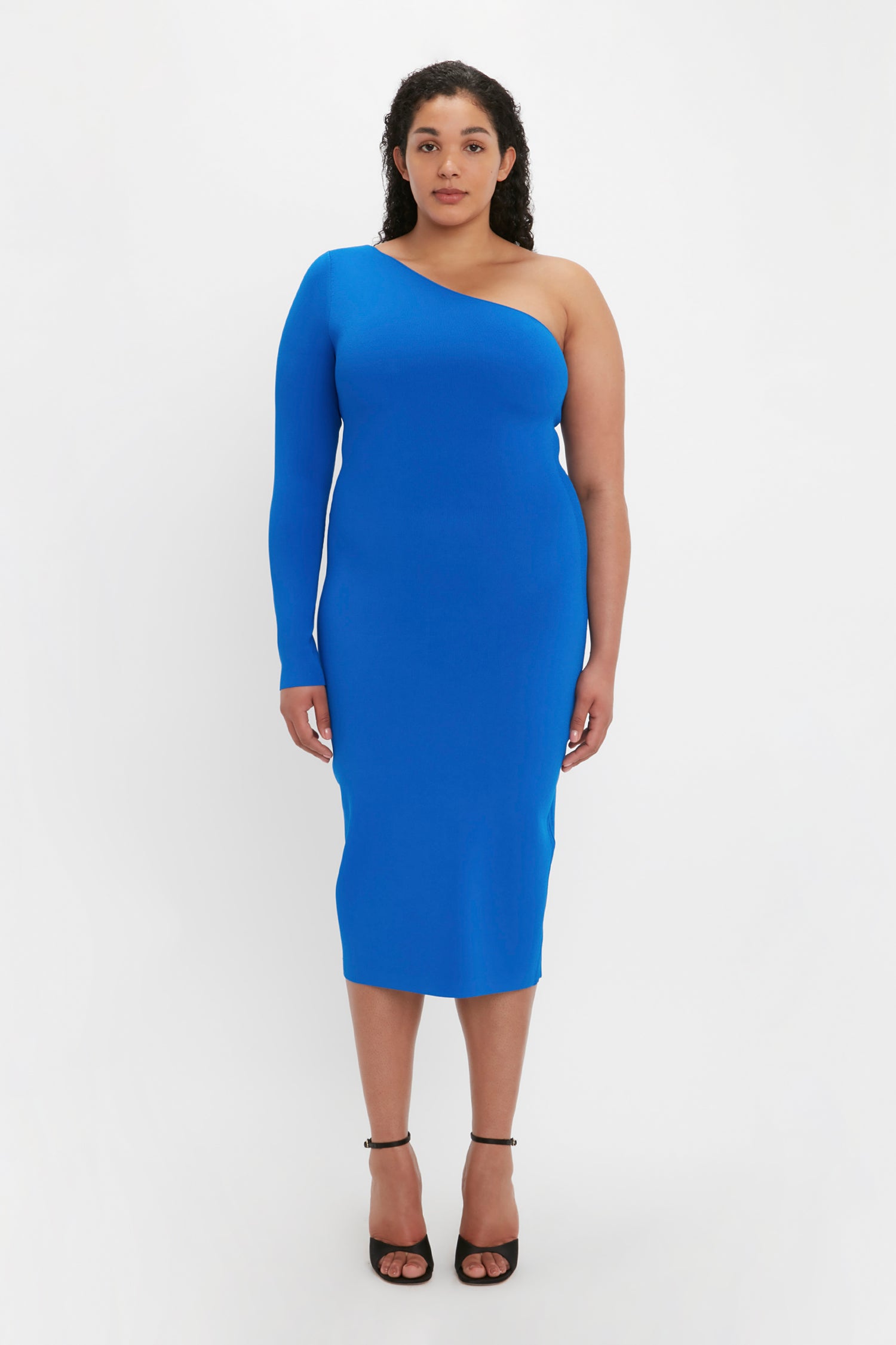 A woman is wearing the Victoria Beckham VB Body One Shoulder Midi Dress in Cobalt, standing against a white background. She has black curly hair and is wearing black high-heeled shoes.