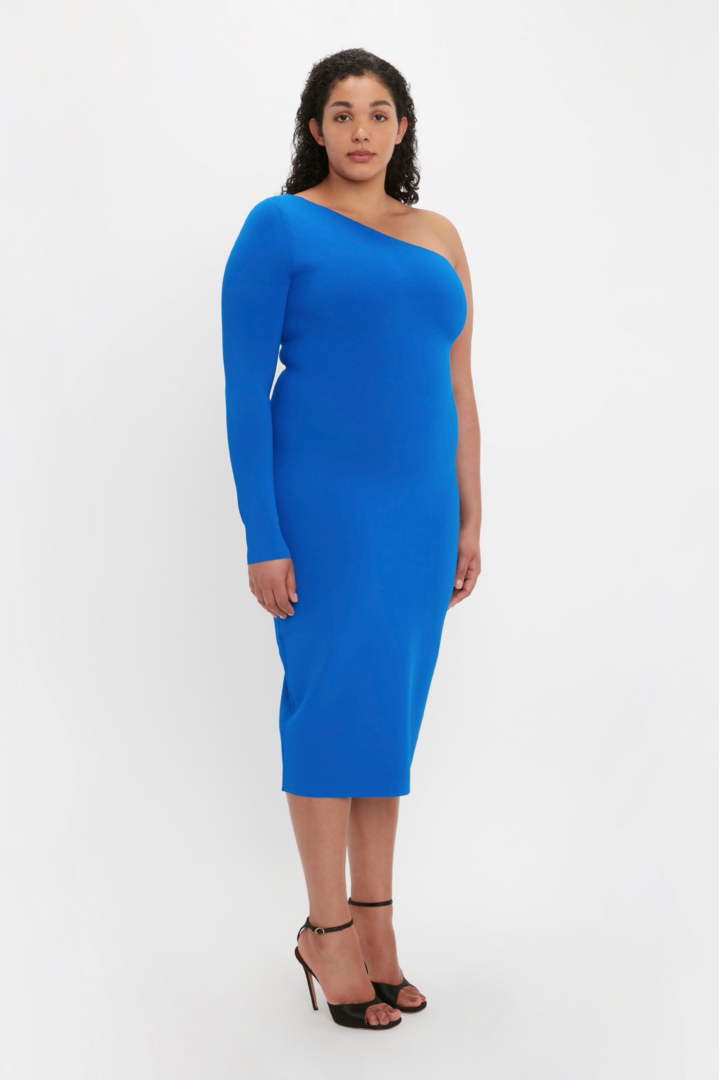 A woman with curly hair is wearing a Victoria Beckham VB Body One Shoulder Midi Dress in Cobalt and black high-heeled shoes, standing against a plain white background.