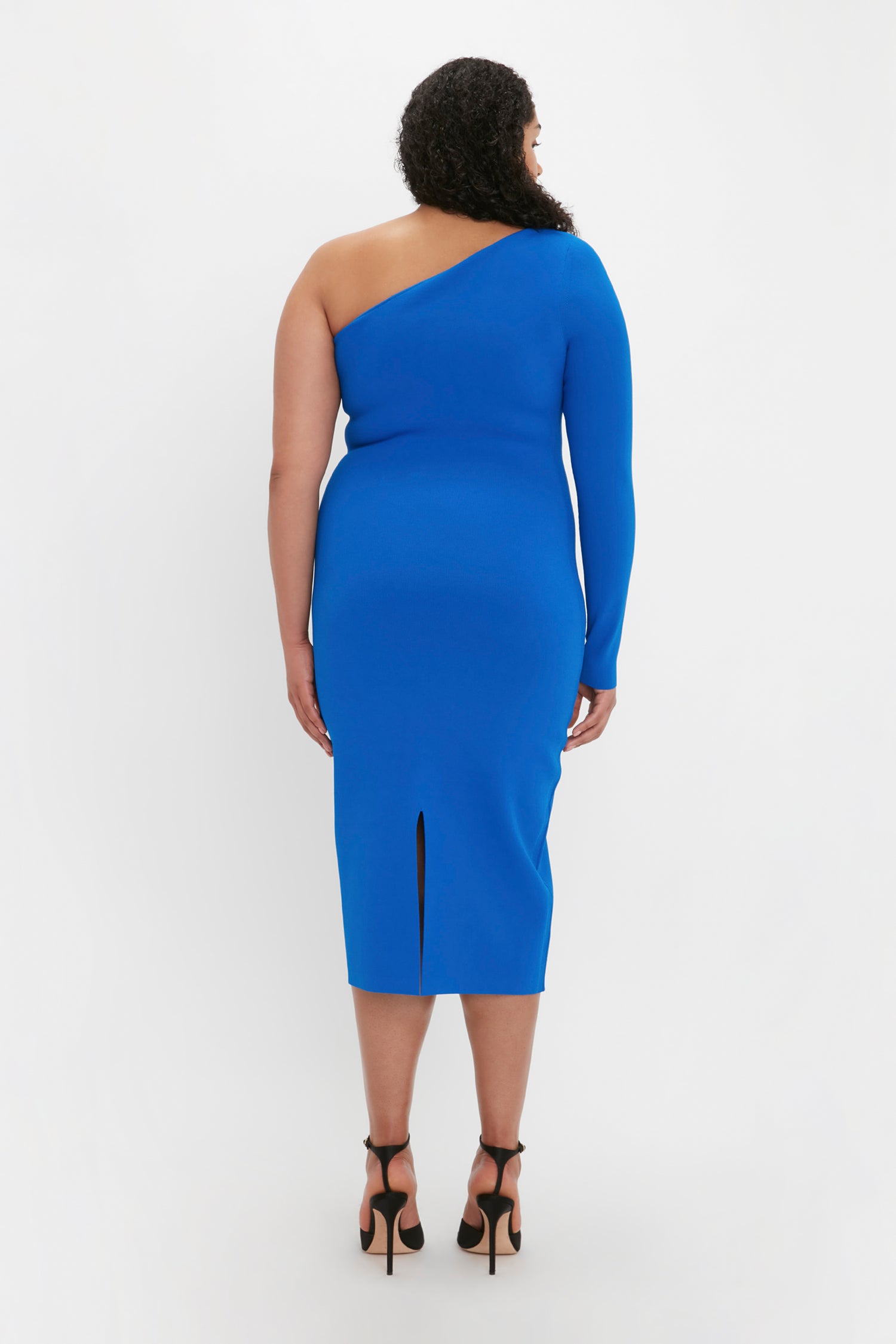 A person with curly hair is standing with their back to the camera, wearing the Victoria Beckham VB Body One Shoulder Midi Dress in Cobalt and black high-heeled sandals. The dress features a slit at the back.