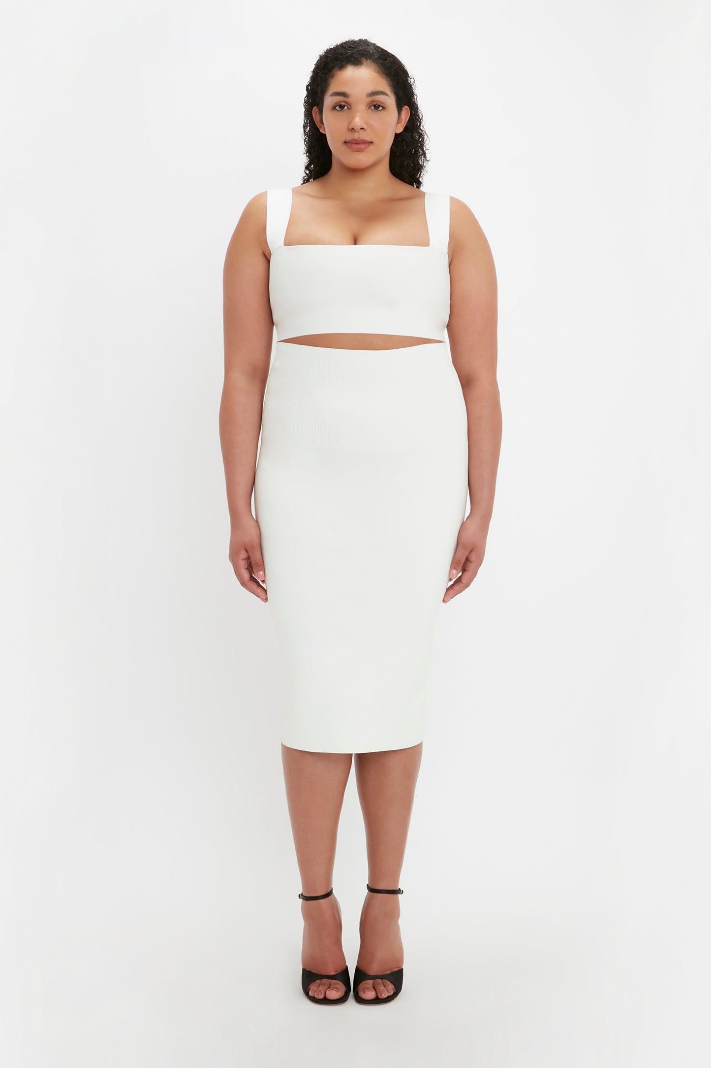 A woman stands against a plain white background wearing the VB Body Strap Bandeau Top in White by Victoria Beckham, paired with a white sleeveless dress featuring a midriff cutout and black high heels.