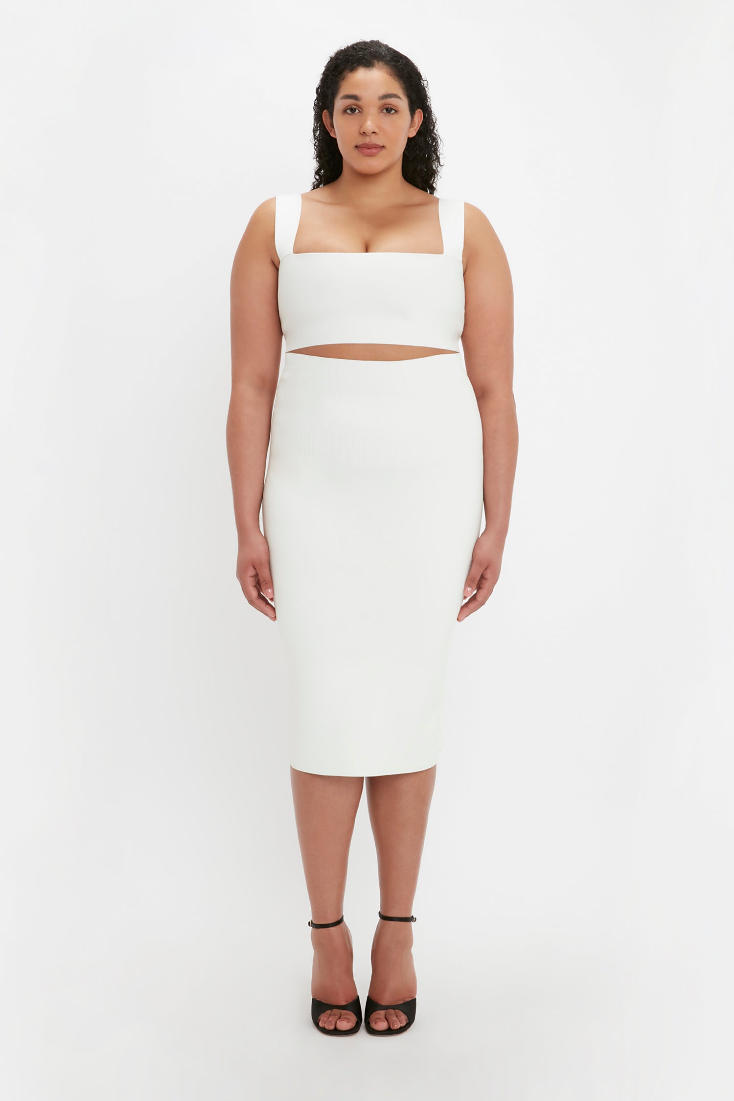 A woman stands against a plain white background wearing the VB Body Strap Bandeau Top in White by Victoria Beckham, paired with a white sleeveless dress featuring a midriff cutout and black high heels.