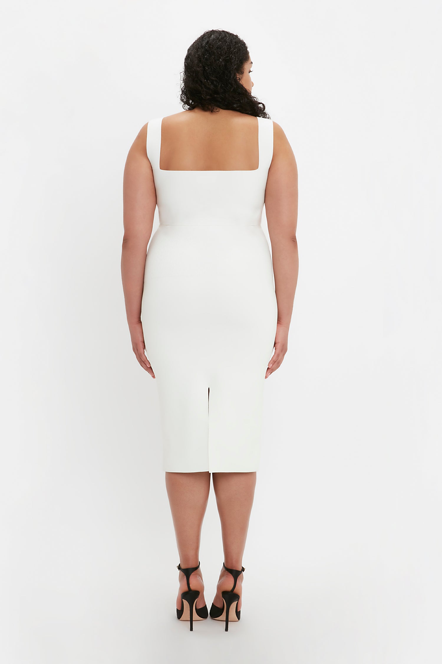A woman with curly hair is standing with her back facing the camera, wearing a sleeveless, square-neck dress and black high-heeled sandals, featuring the VB Body Strap Bandeau Top in White by Victoria Beckham.