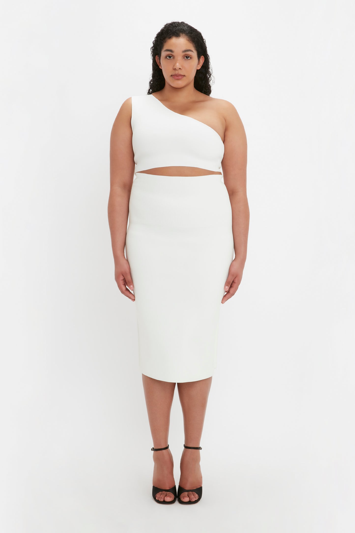 A woman stands facing forward, wearing the VB Body Fitted Midi Skirt in white by Victoria Beckham and black high heels, with a neutral expression against a white background.