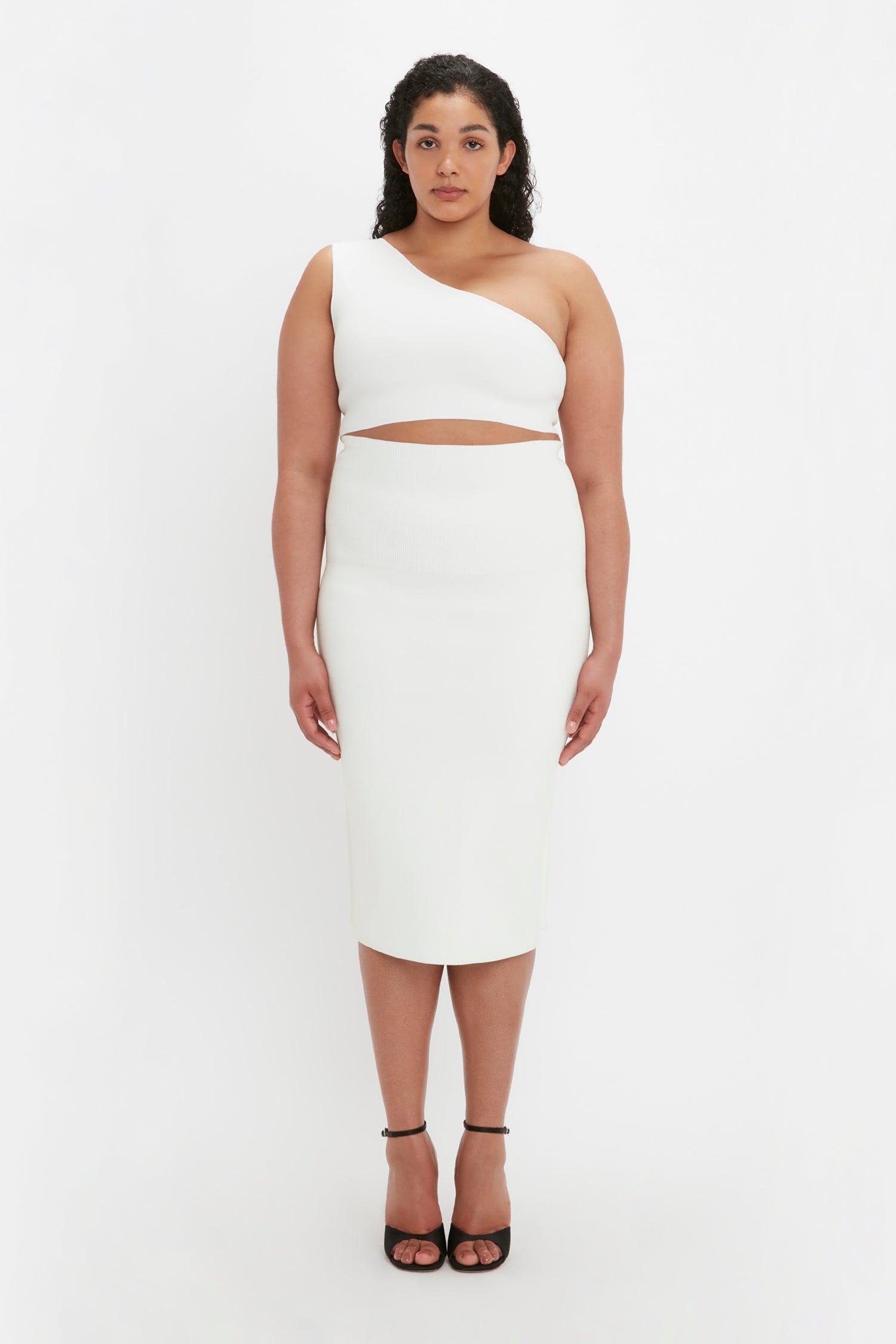 A woman stands facing forward, wearing the VB Body Fitted Midi Skirt in white by Victoria Beckham and black high heels, with a neutral expression against a white background.