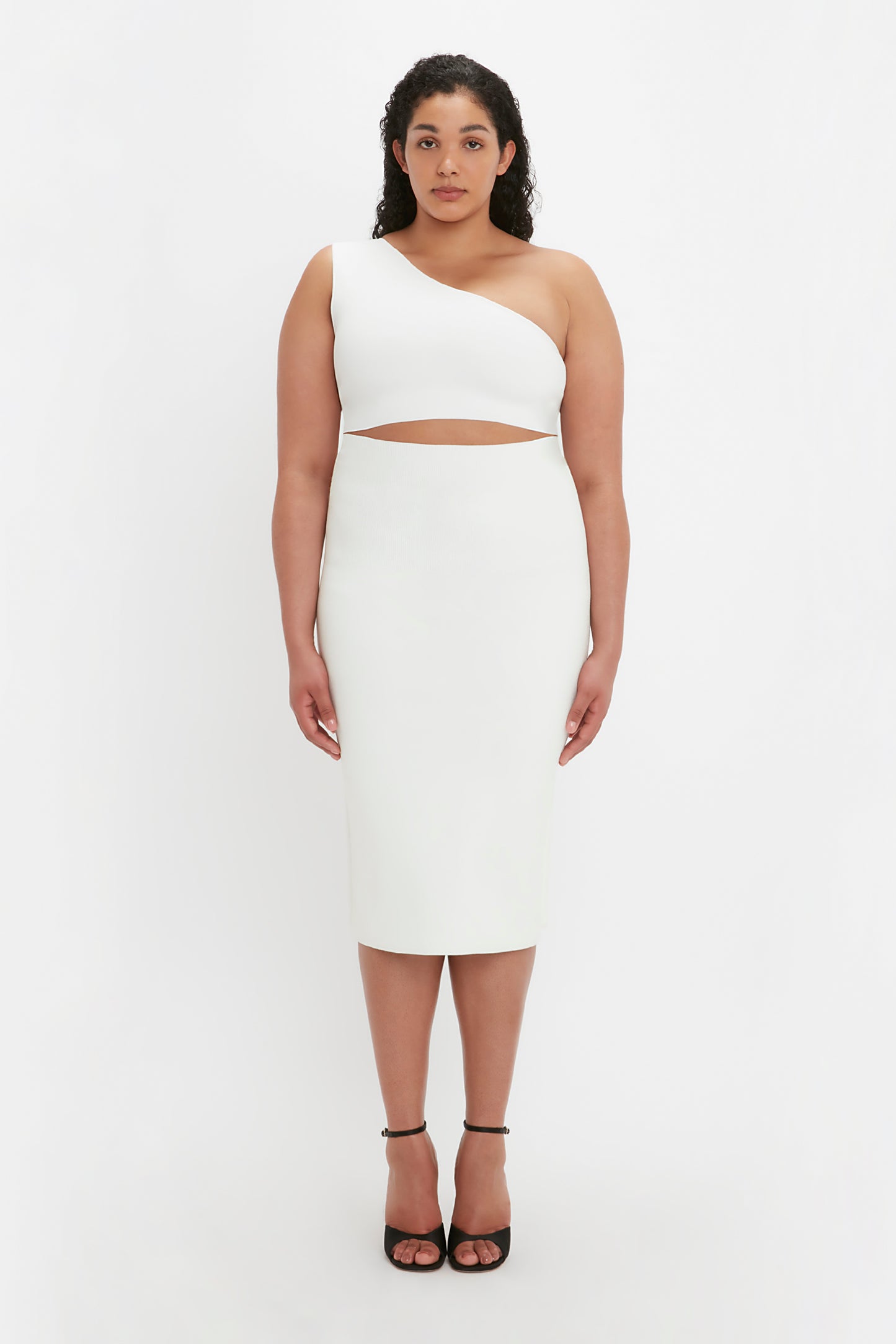 A woman wearing the VB Body One Shoulder Crop Top in white from Victoria Beckham stands against a plain white background. She has long, curly hair and is wearing black high-heeled shoes.