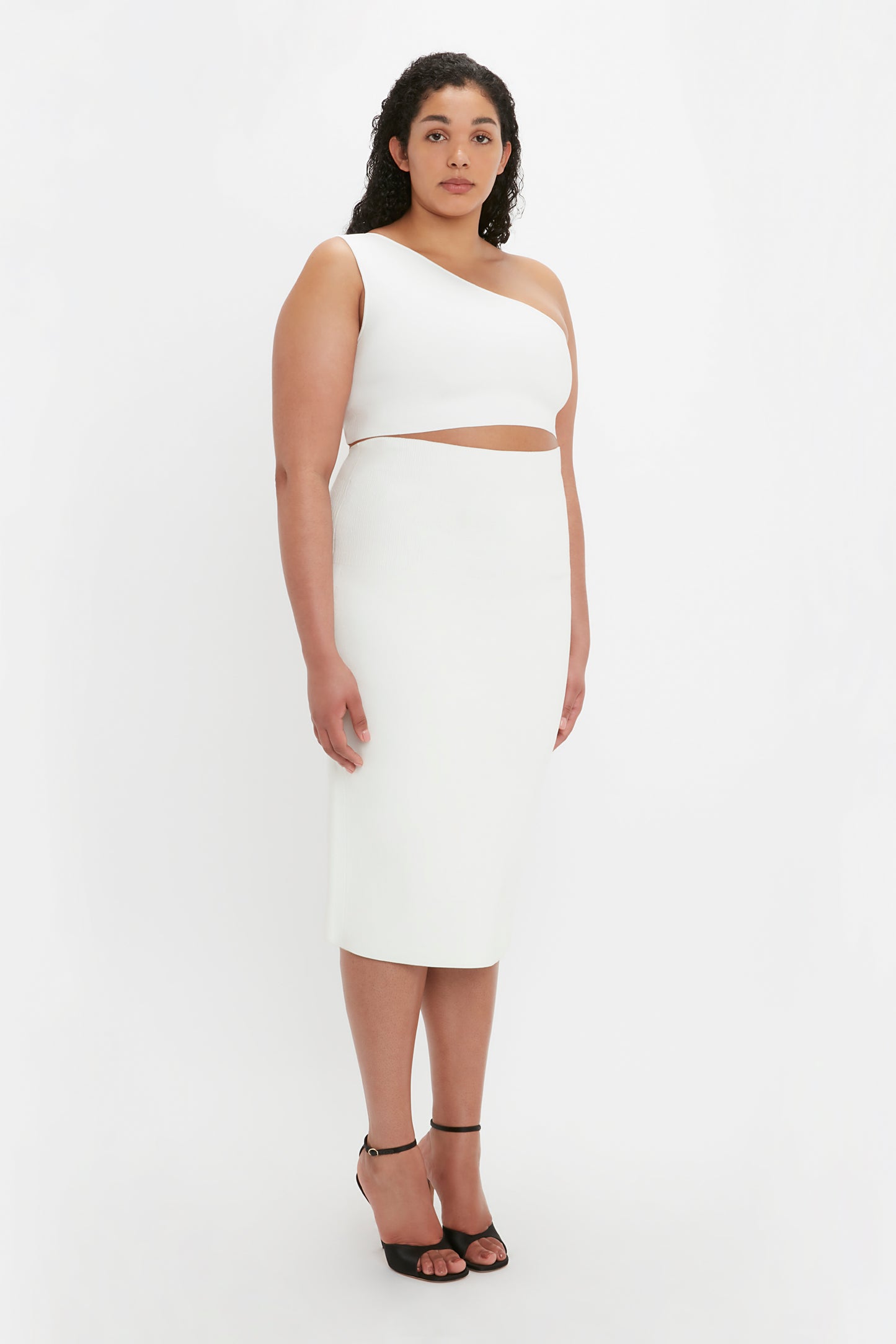 A woman stands against a white background wearing the VB Body Fitted Midi Skirt in White by Victoria Beckham and black high-heeled sandals. She has curly hair and is looking at the camera.