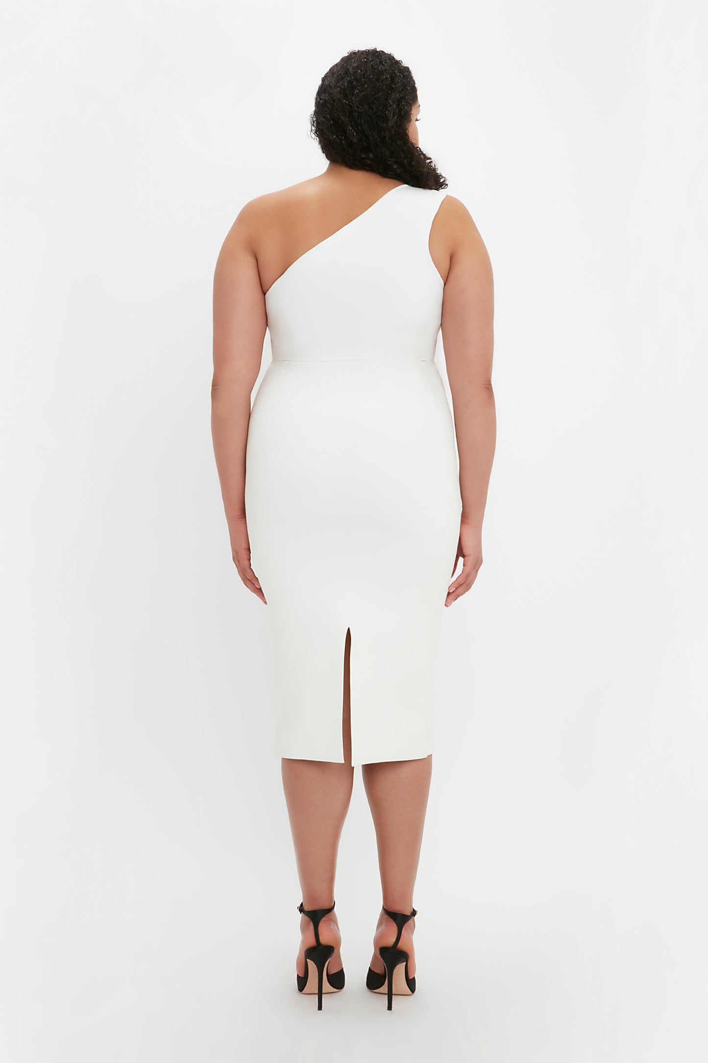 A person with curly hair wearing a white VB Body One Shoulder Crop Top by Victoria Beckham and black high-heeled sandals is standing facing away from the camera.