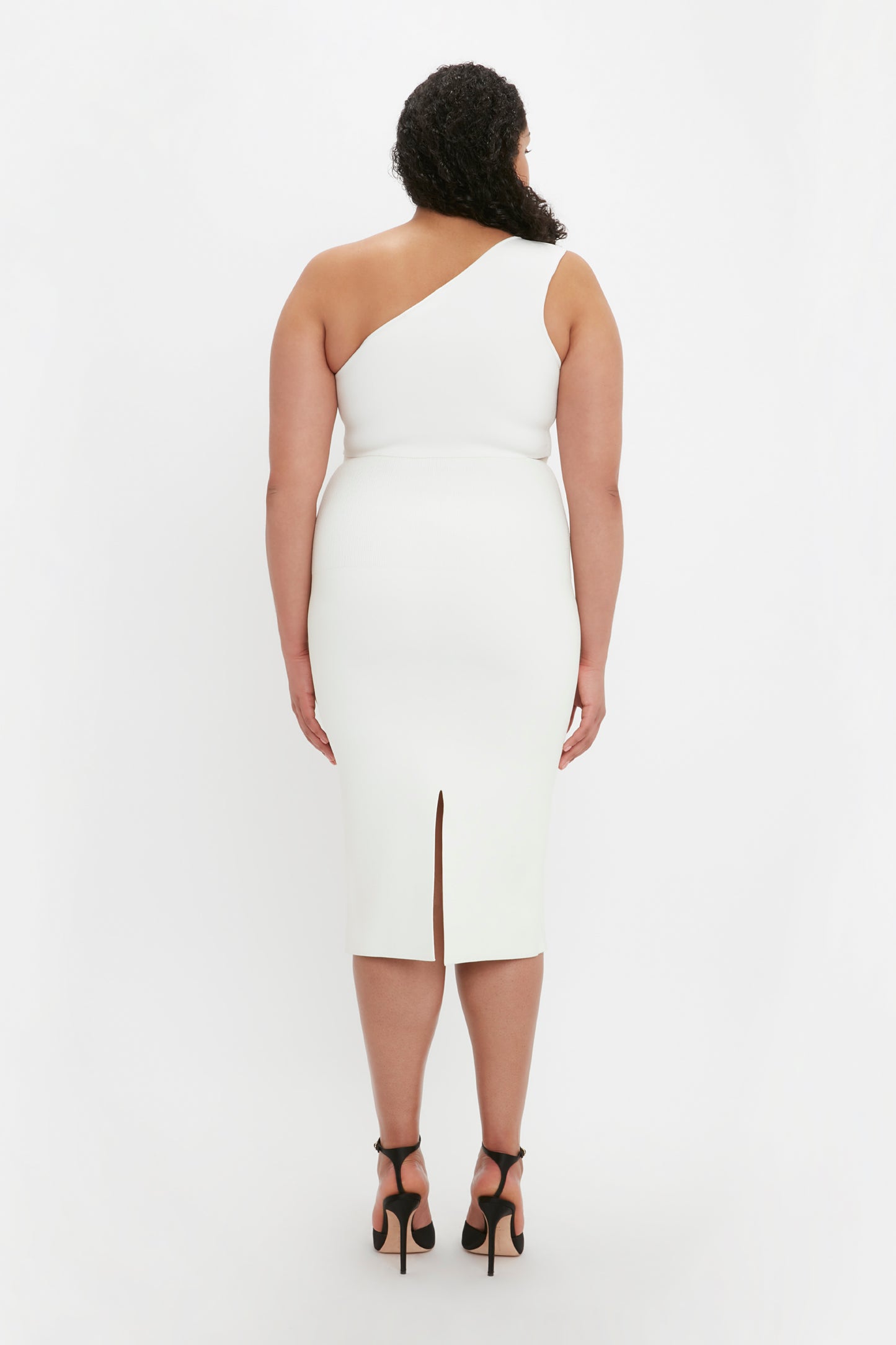 A woman with curly hair is seen from the back, wearing the VB Body Fitted Midi Skirt in white by Victoria Beckham along with a sleeveless, one-shoulder top and black high heels.