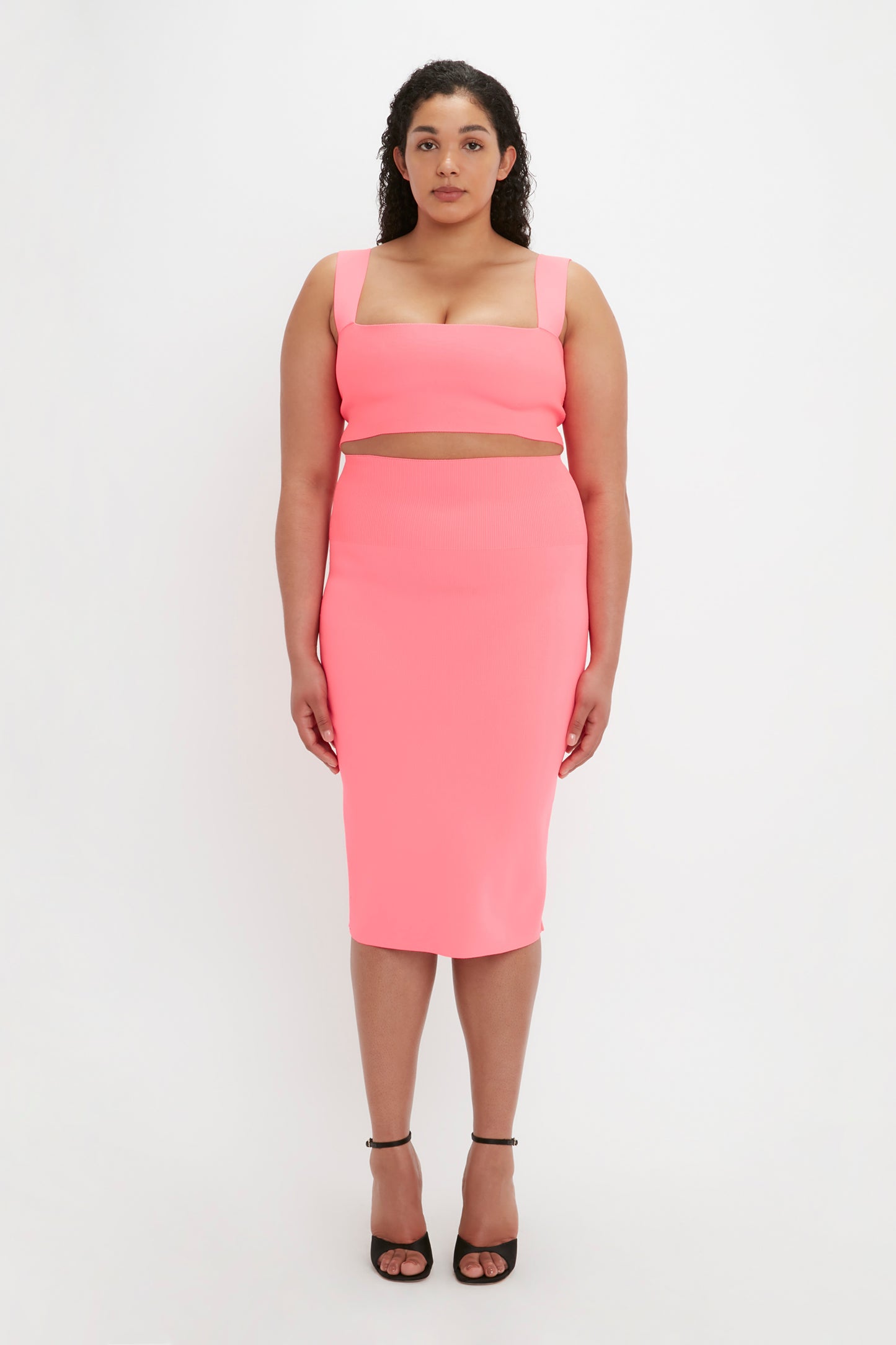 A woman stands facing forward, wearing a pink VB Body Fitted Midi Skirt by Victoria Beckham along with a matching crop top. She has long, curly hair and is sporting black high heels against a plain white background.