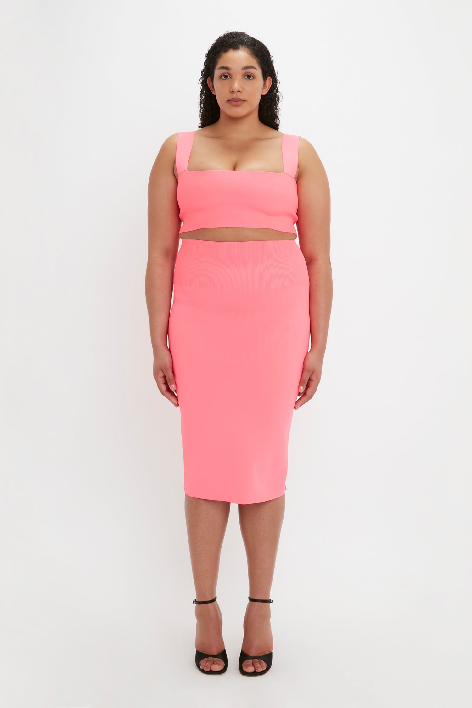 A woman stands facing forward, wearing a pink VB Body Fitted Midi Skirt by Victoria Beckham along with a matching crop top. She has long, curly hair and is sporting black high heels against a plain white background.