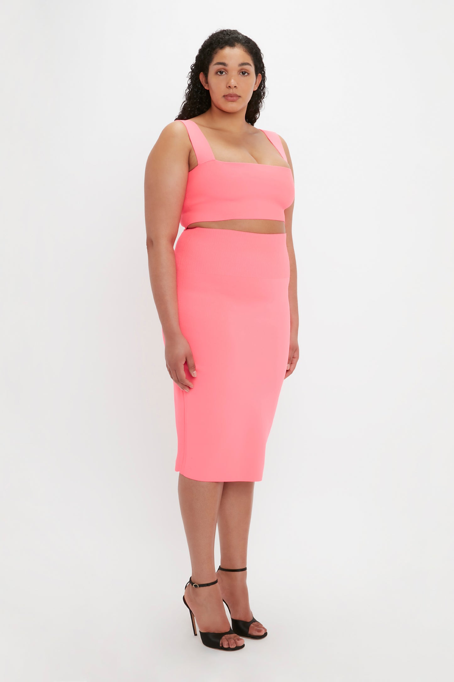 A woman with curly hair is wearing the Victoria Beckham VB Body Fitted Midi Skirt in Pink paired with a matching neon pink crop top and black high-heeled sandals, standing against a plain white background.
