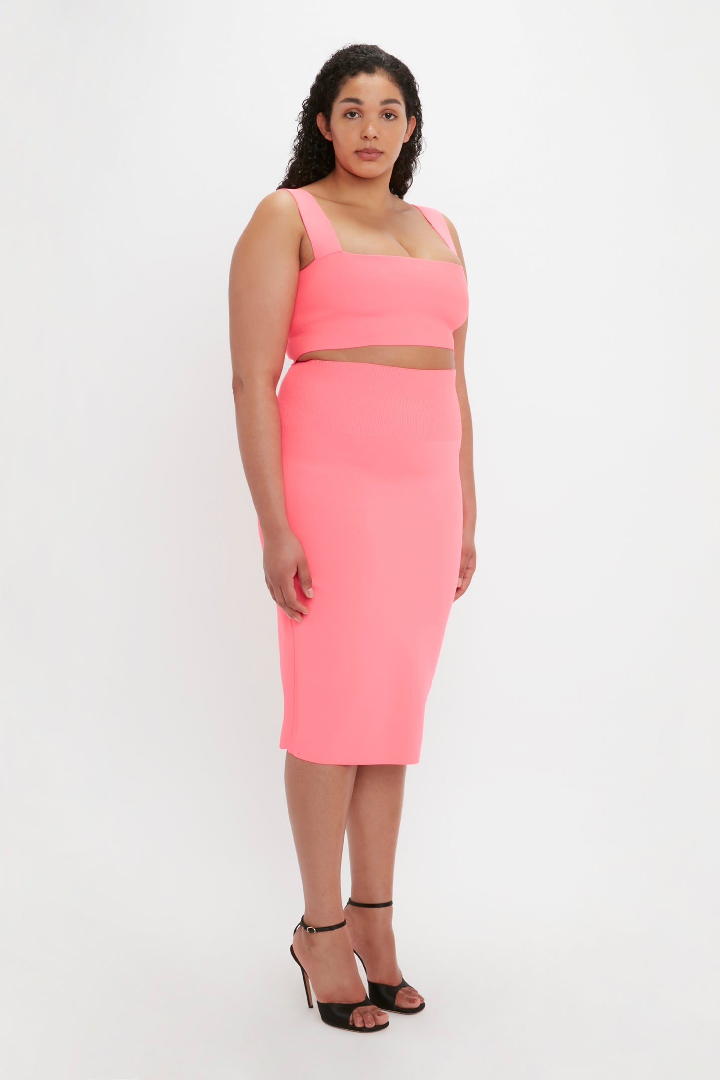 A woman with curly hair is wearing the Victoria Beckham VB Body Strap Bandeau Top in Pink, accompanied by a matching pink skirt and black heels, standing against a plain white background.