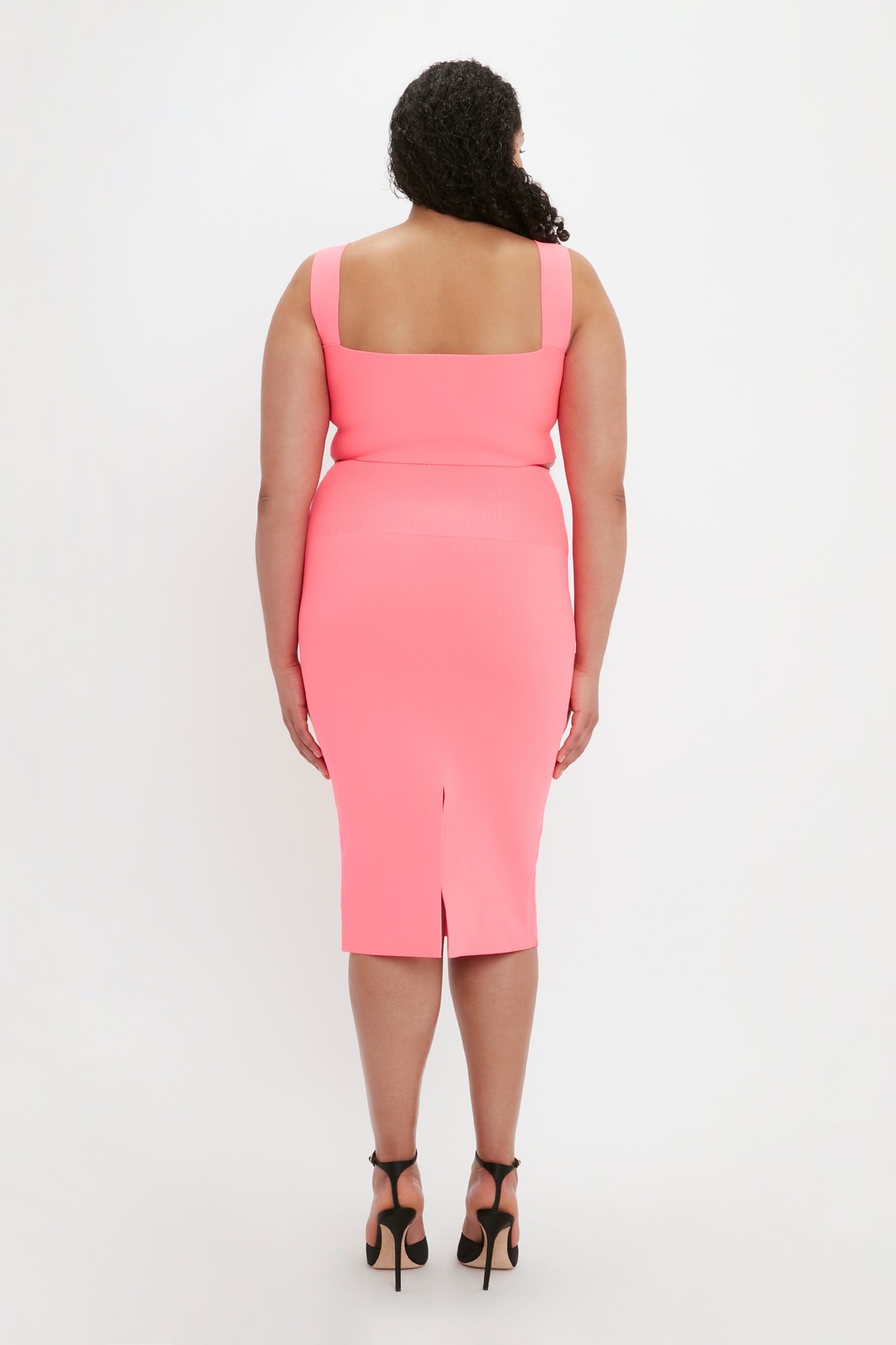 A woman is standing with her back facing the camera, wearing the Victoria Beckham VB Body Fitted Midi Skirt in Pink and black high-heeled shoes.
