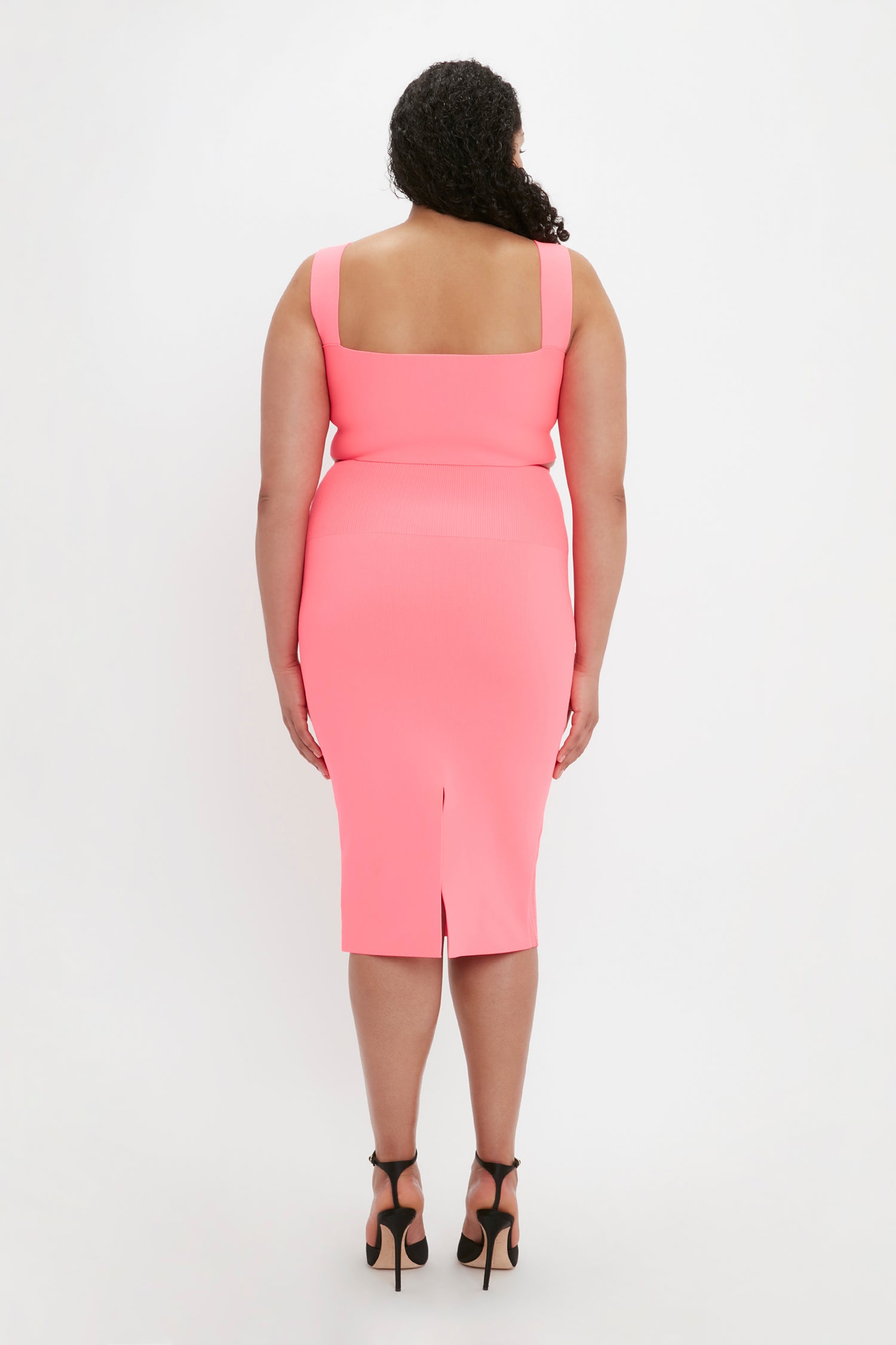 A woman is standing with her back facing the camera, wearing the Victoria Beckham VB Body Fitted Midi Skirt in Pink and black high-heeled shoes.