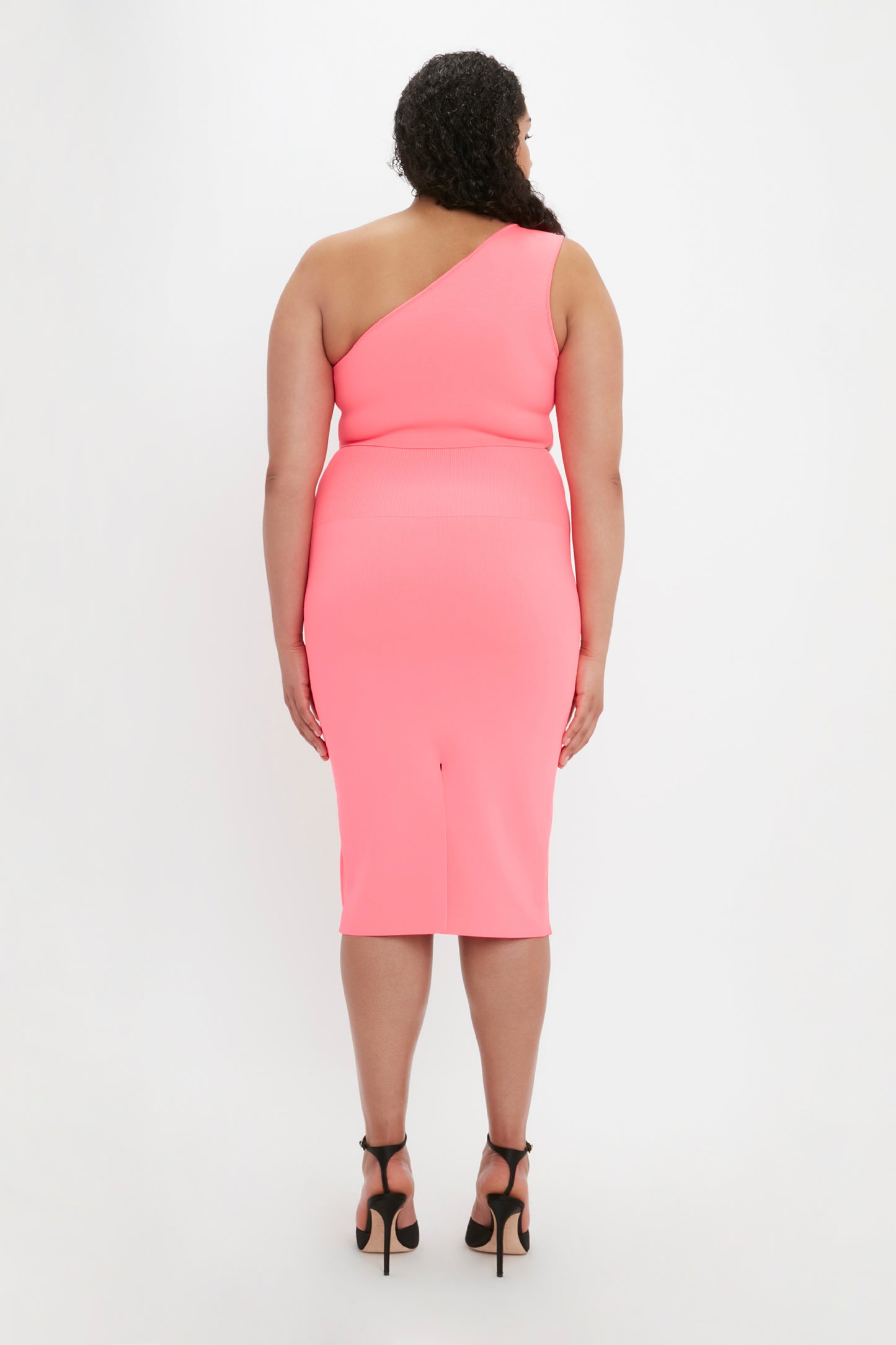 Person wearing a VB Body One Shoulder Crop Top in Pink by Victoria Beckham and black high heels, facing away from the camera against a plain white background.