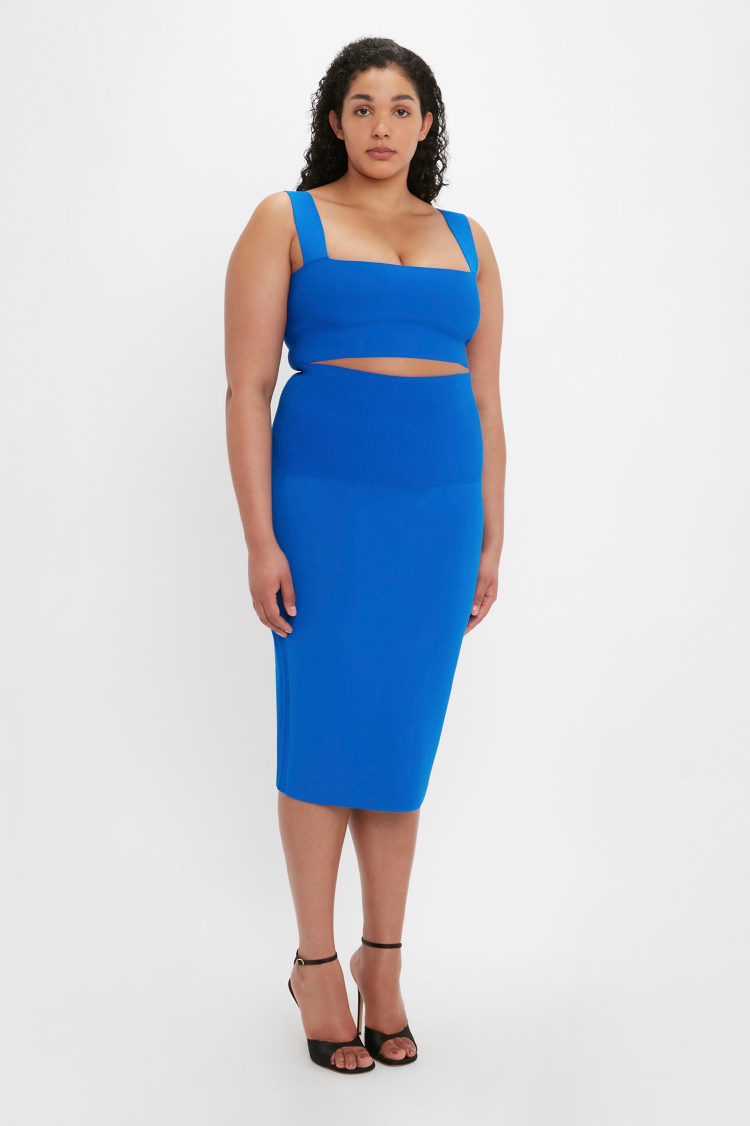 A woman with curly hair stands against a white background, wearing a blue sleeveless crop top, the Victoria Beckham VB Body Fitted Midi Skirt in cobalt, and black heeled sandals.
