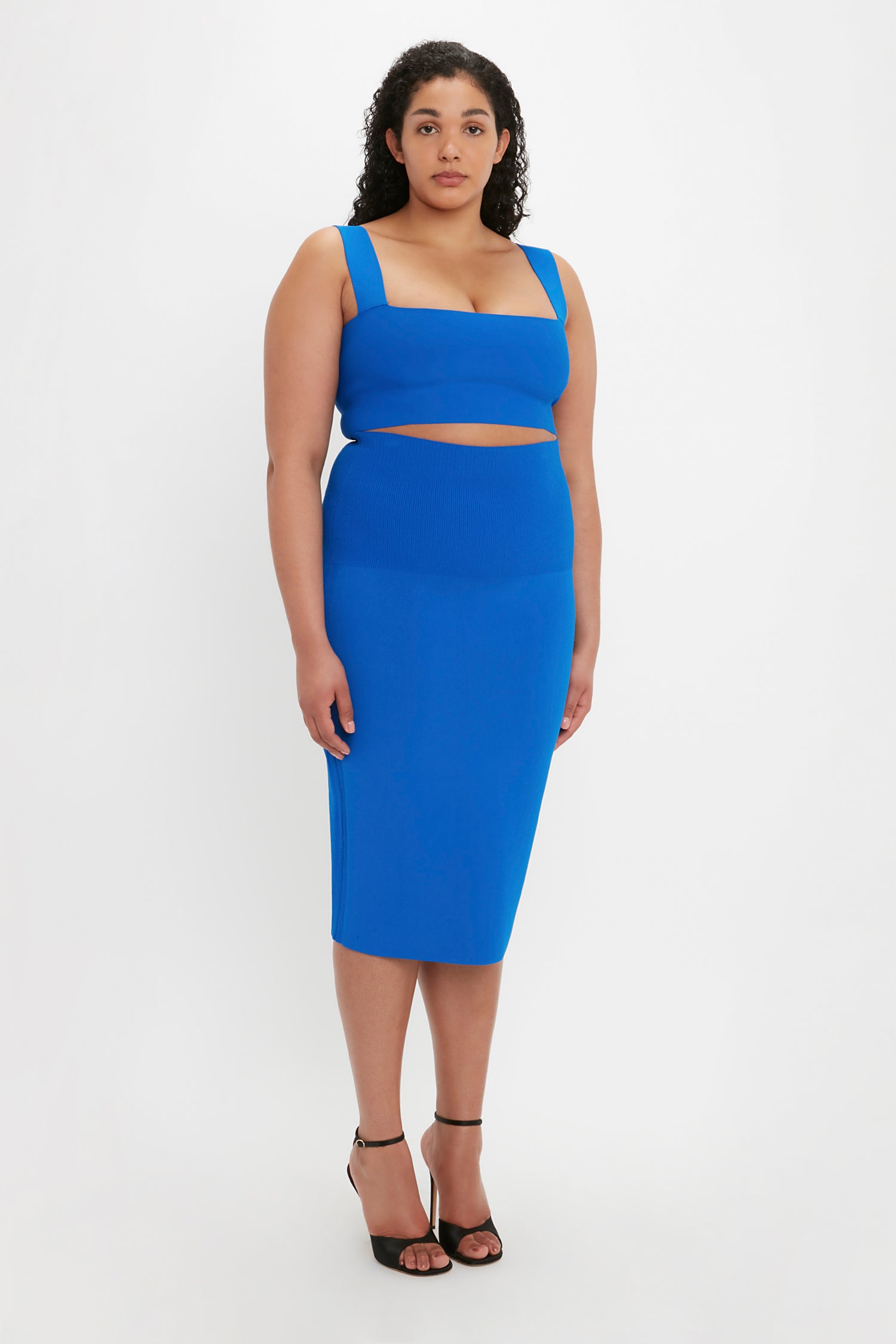 A person with curly hair stands against a plain white background, wearing the Victoria Beckham VB Body Strap Bandeau Top in Cobalt paired with matching blue bottoms and black heels.