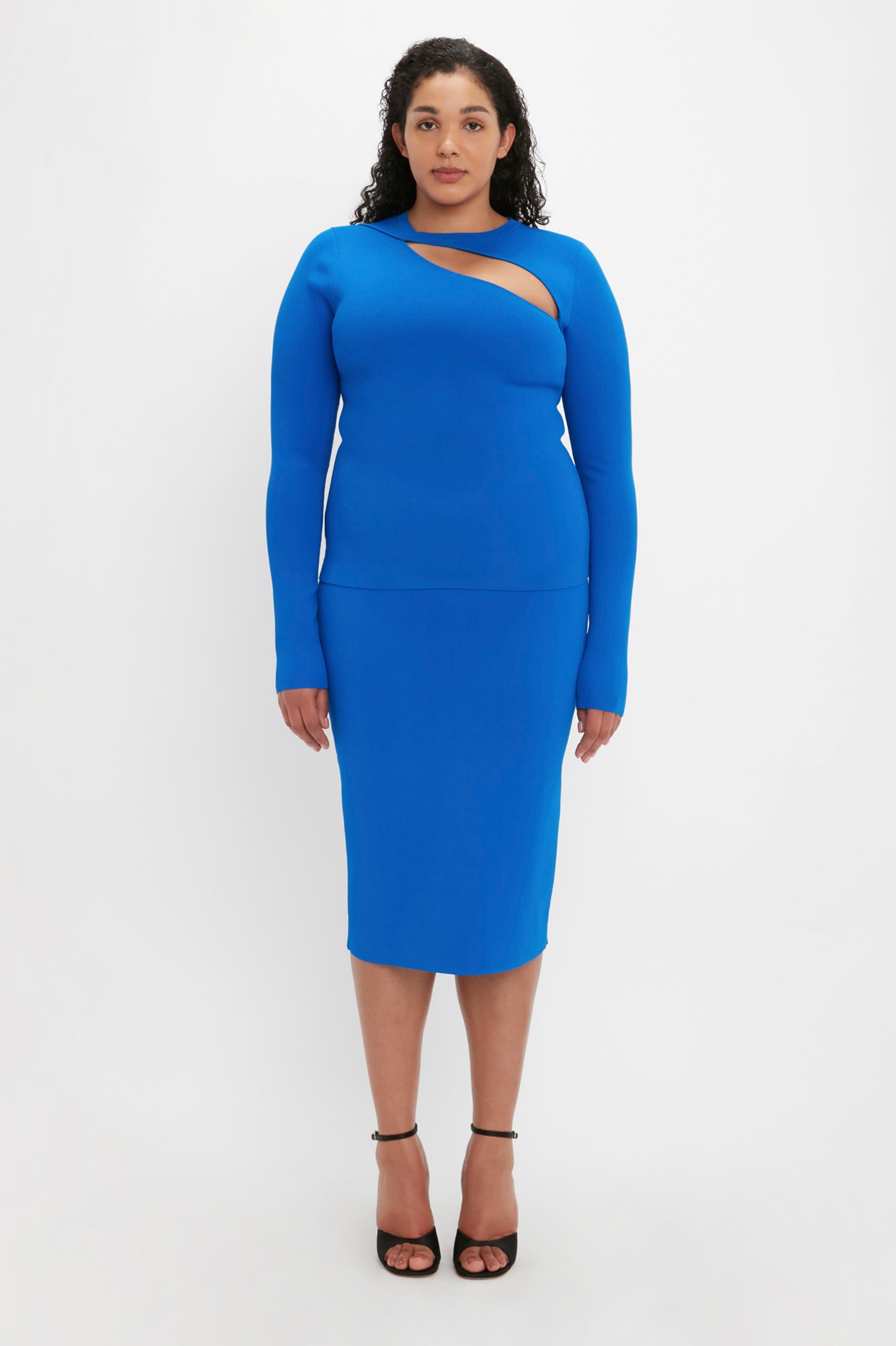 A person stands against a white background wearing the Victoria Beckham VB Body Front Slash Crew Neck Top in Cobalt and black high-heeled sandals.