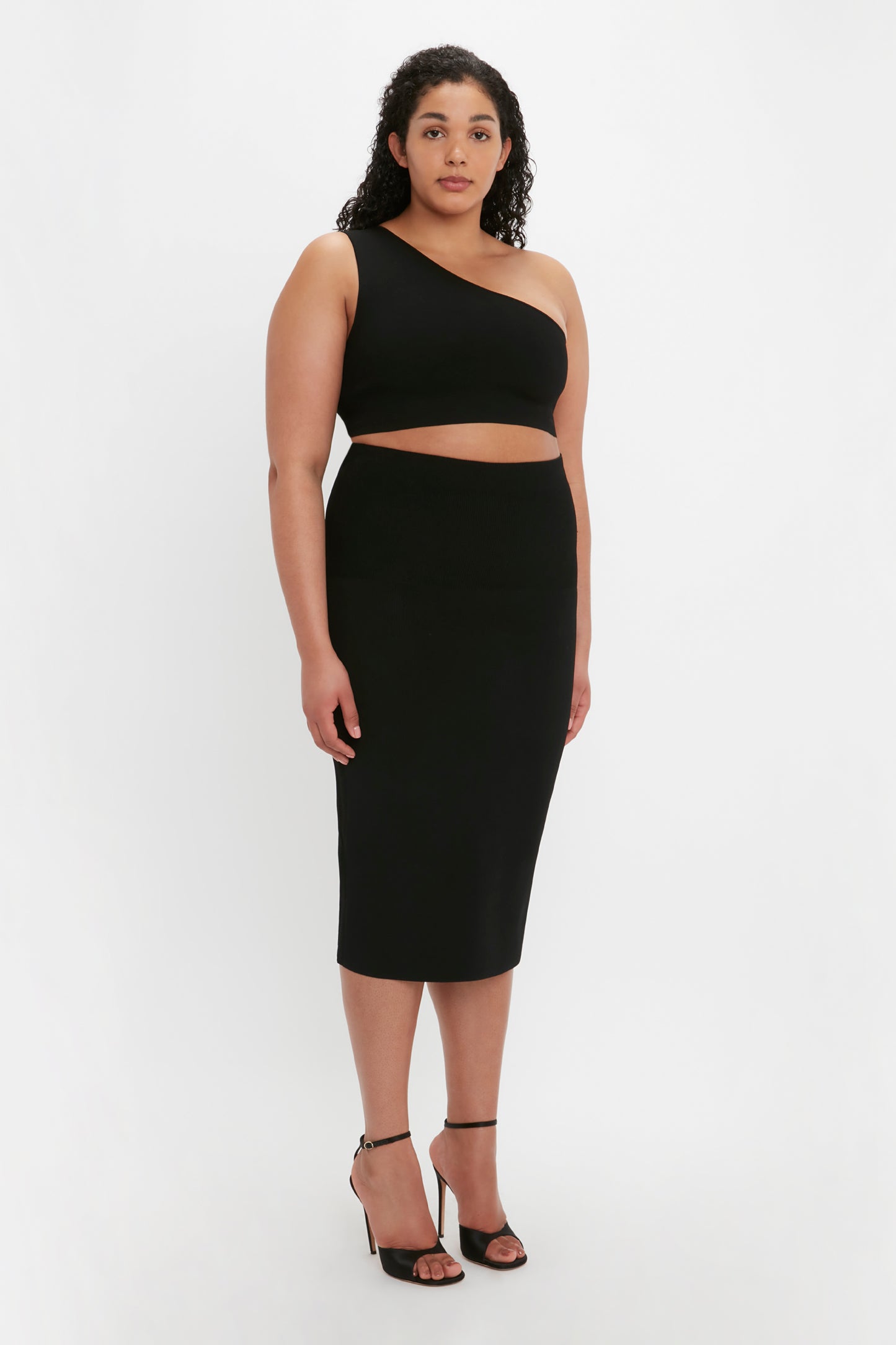 A person with curly hair is wearing a one-shoulder black crop top, the VB Body Fitted Midi Skirt in Black by Victoria Beckham, and black high-heeled sandals. They are standing against a plain white background.
