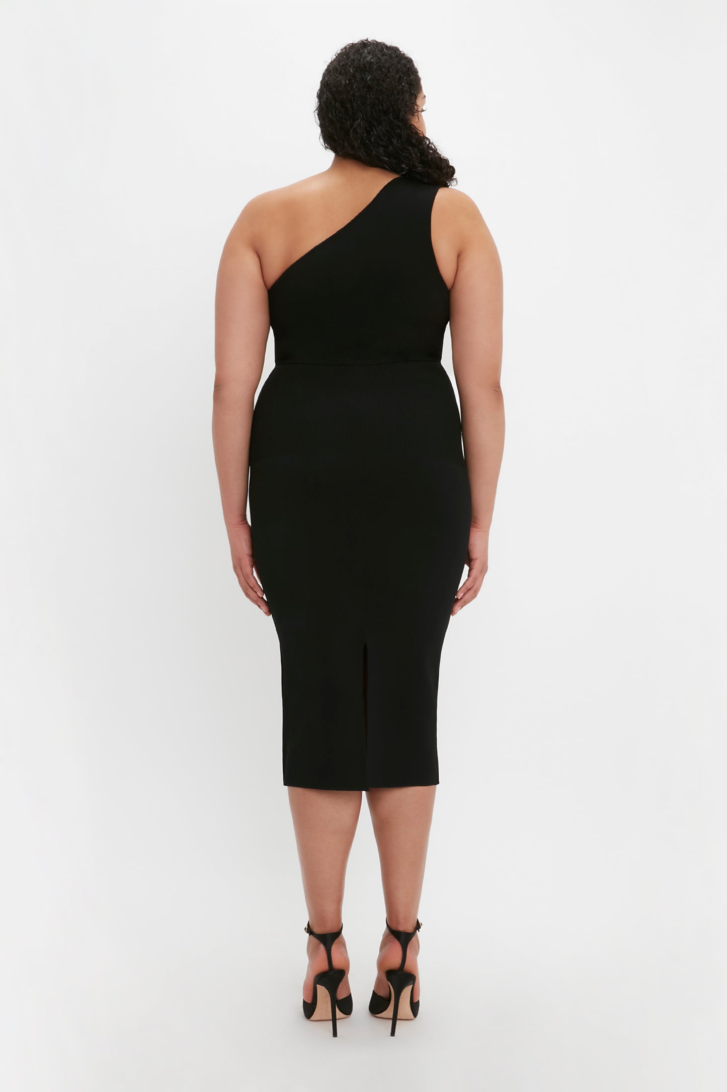 A woman with curly hair is standing and facing away, wearing the VB Body One Shoulder Crop Top in Black by Victoria Beckham and black high heels. The background is plain white.
