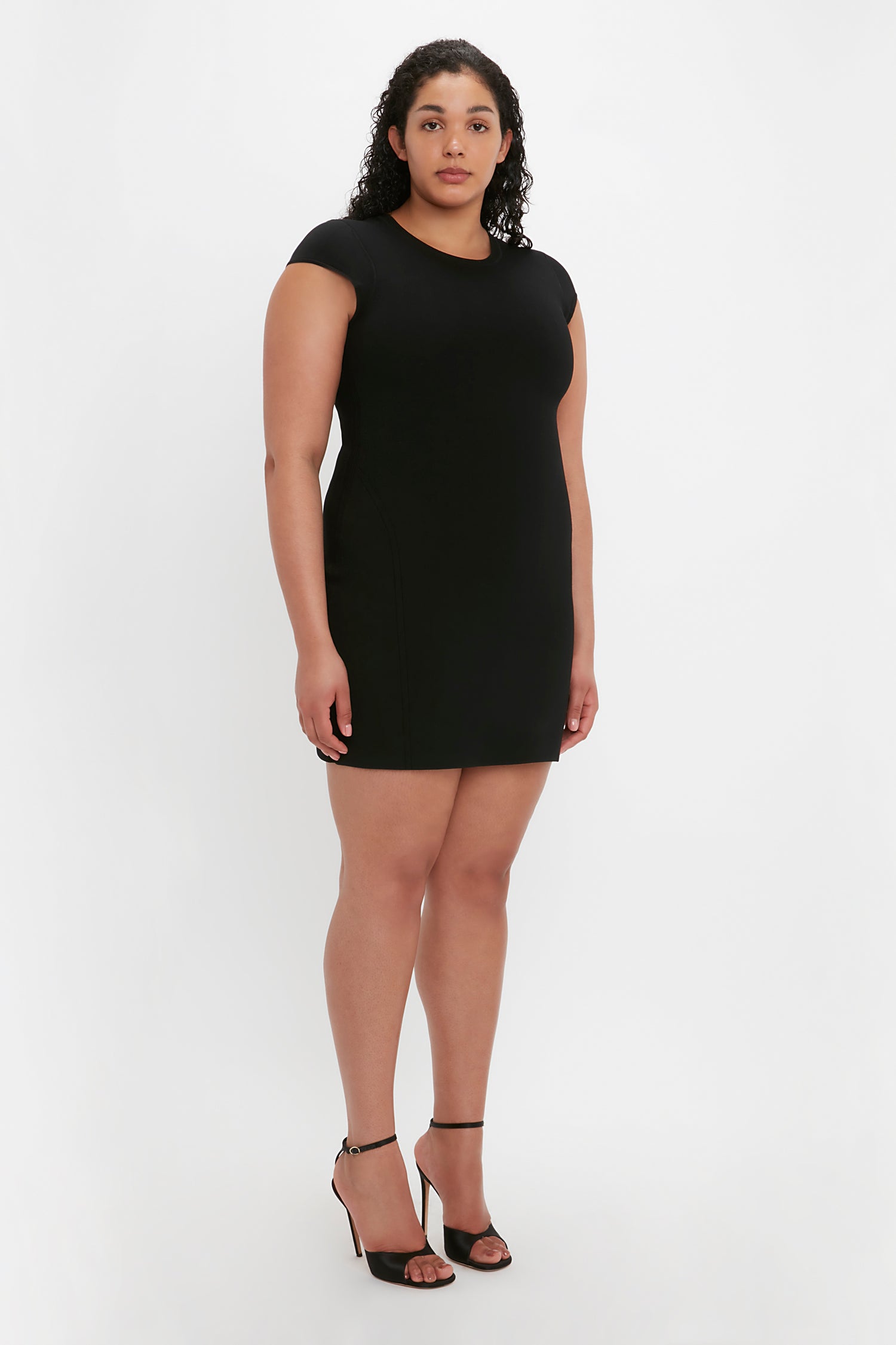 A person with curly hair wearing the VB Body Compact Cap Sleeve Mini Dress in Black by Victoria Beckham and black high-heeled sandals stands against a plain white background.