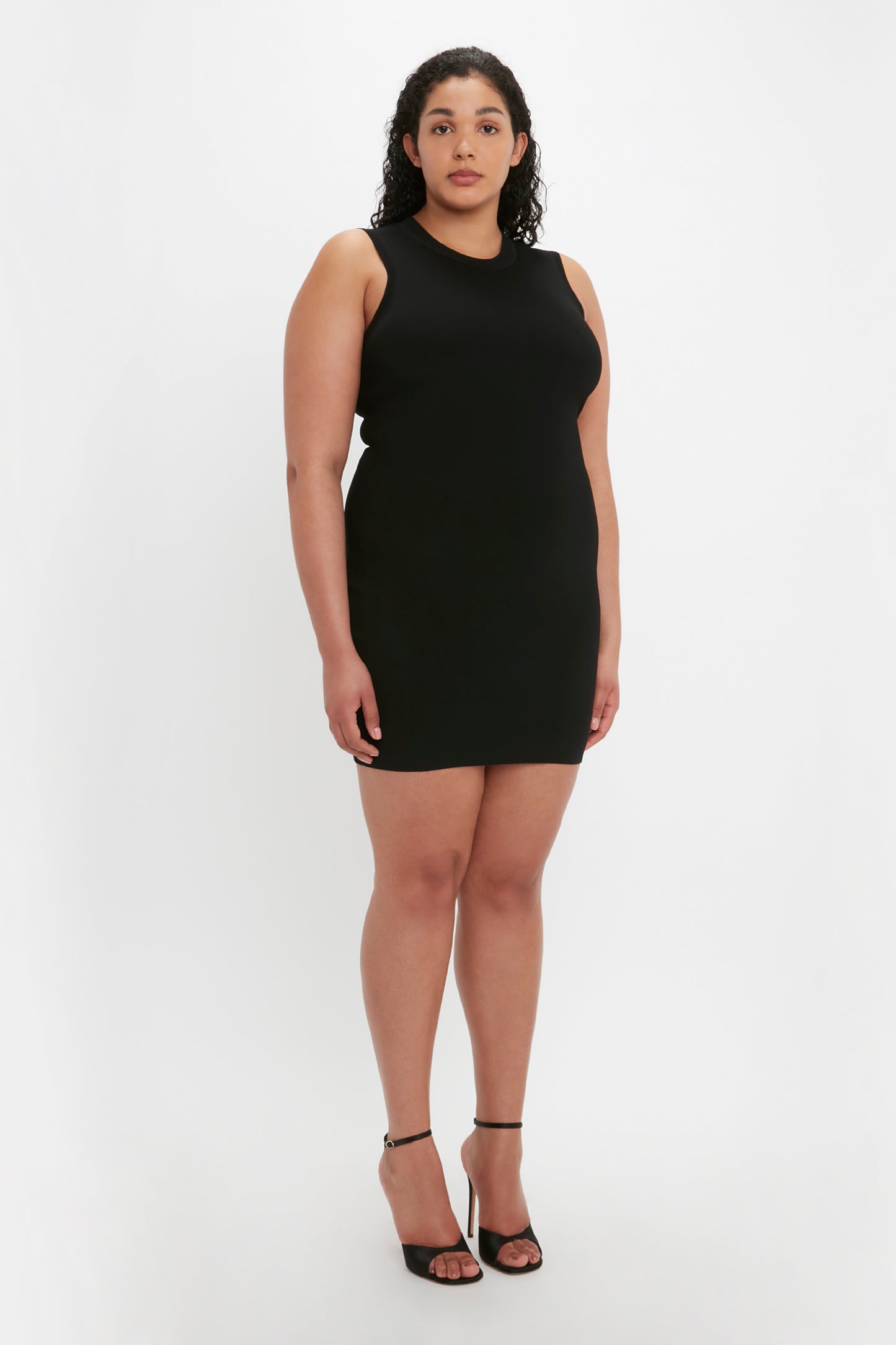 A woman with curly hair is standing and wearing the VB Body Mini Dress in Black by Victoria Beckham, paired with black high-heeled sandals, against a plain white background.