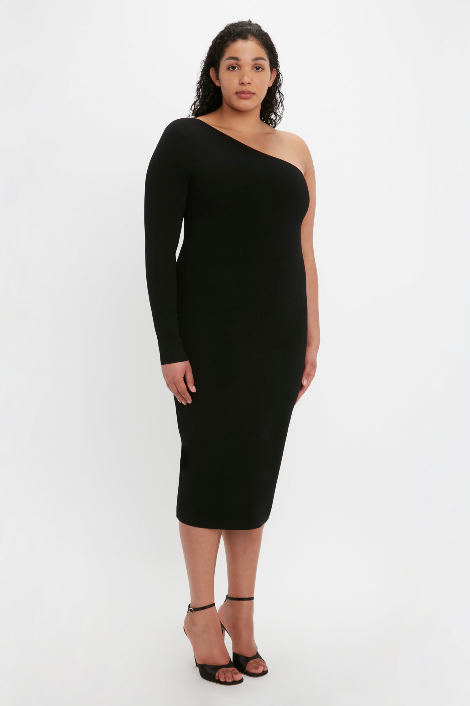 A woman stands against a plain background wearing the Victoria Beckham VB Body One Shoulder Midi Dress in Black paired with black heels.