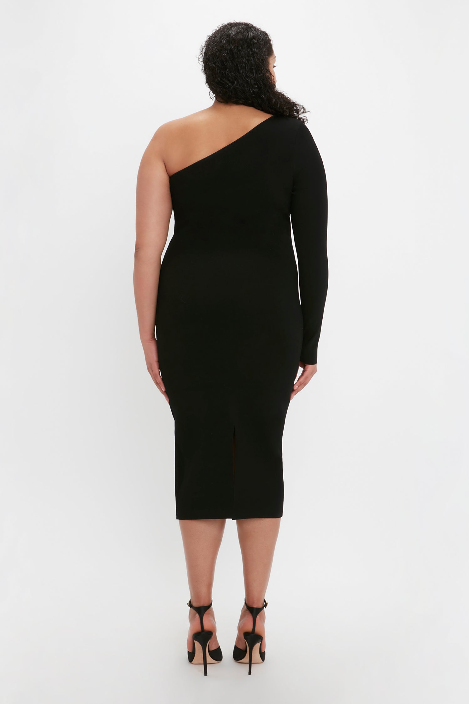 A person with curly hair stands facing away, wearing the VB Body One Shoulder Midi Dress in Black by Victoria Beckham and high-heeled shoes.