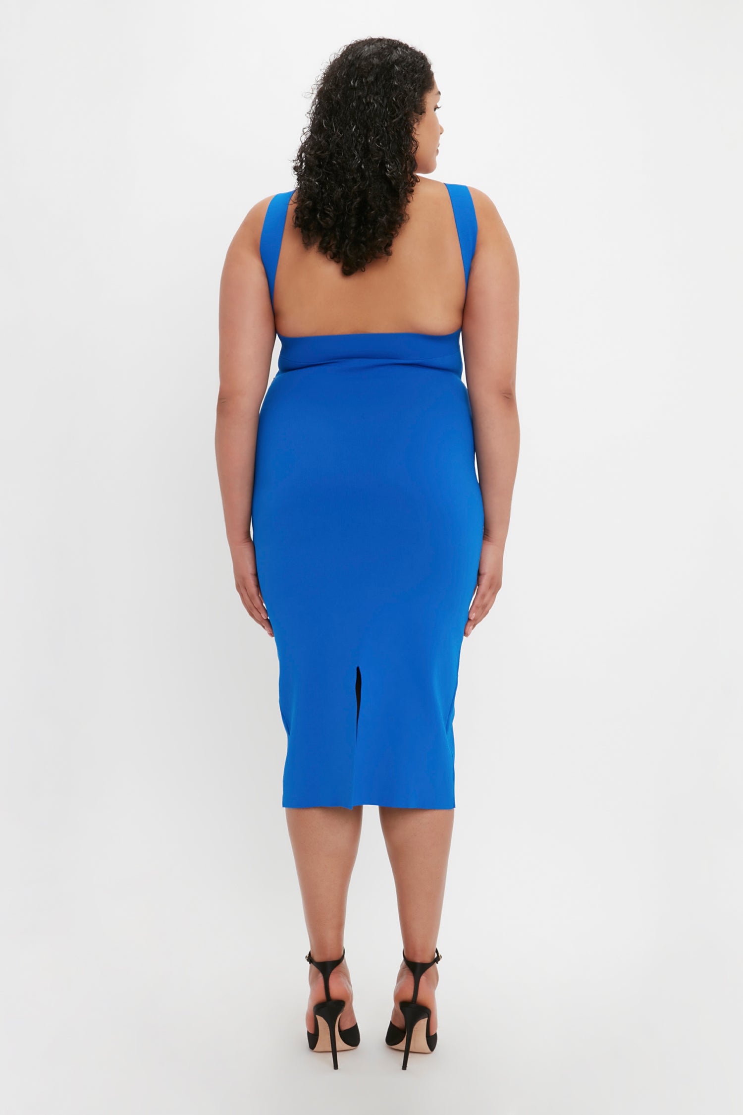 A person with curly hair is standing and facing away, wearing the VB Body Cut Out Midi Dress in Cobalt by Victoria Beckham and black high heels against a plain white background.