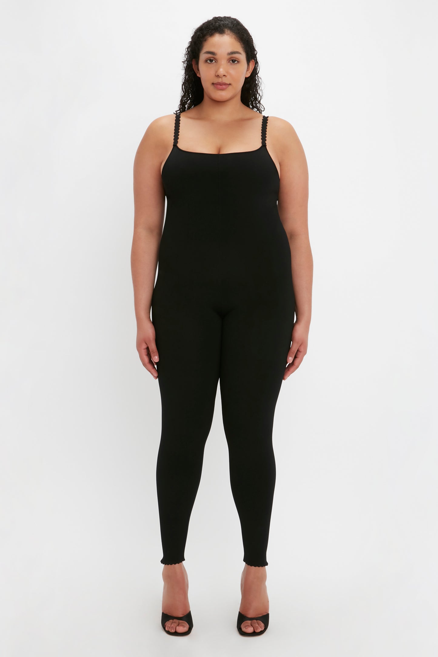 A woman stands facing forward, wearing the VB Body Catsuit in Black by Victoria Beckham and black high-heeled shoes. She has curly hair and a neutral expression, set against a plain white background.