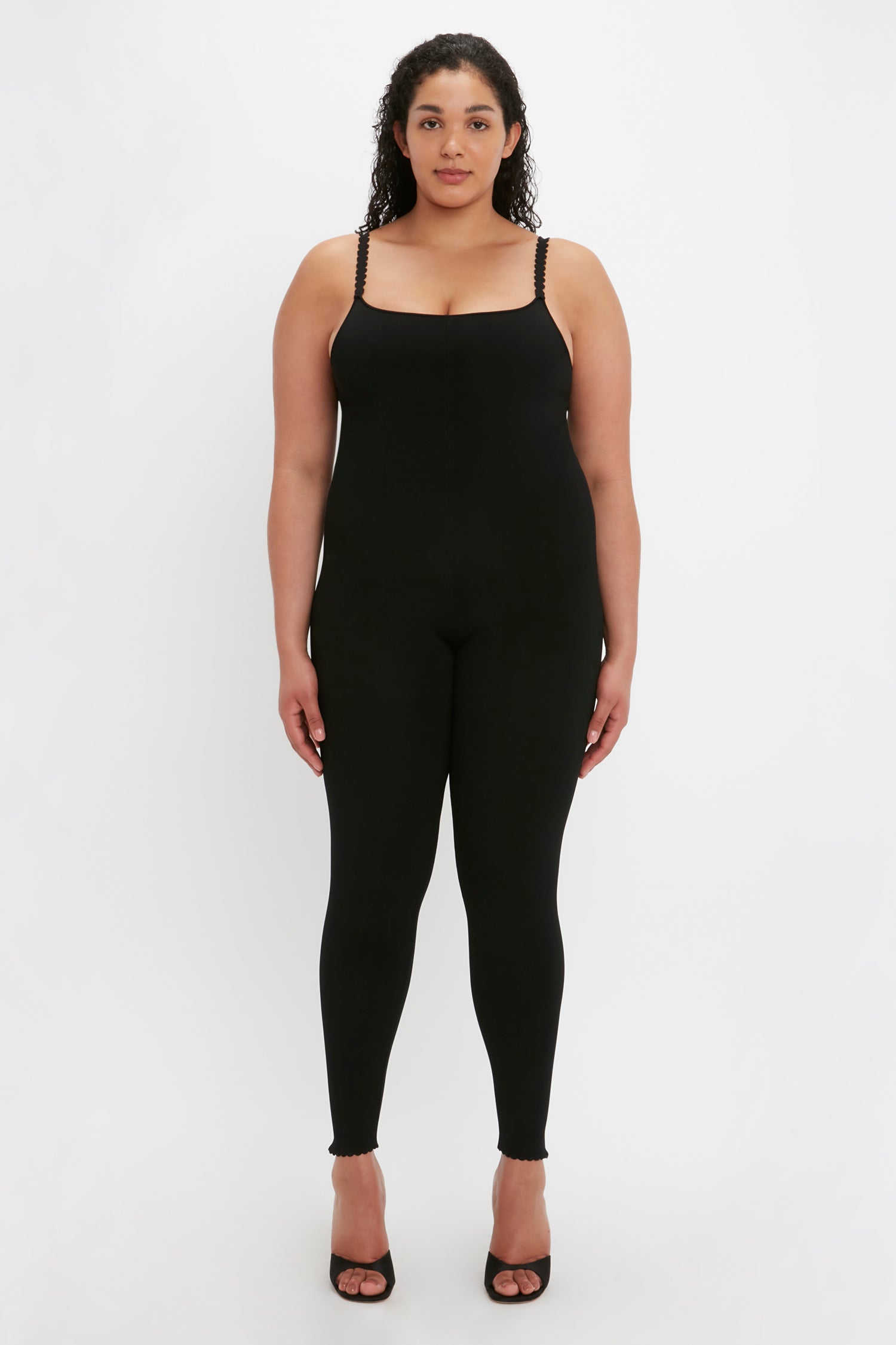 A woman stands facing forward, wearing the VB Body Catsuit in Black by Victoria Beckham and black high-heeled shoes. She has curly hair and a neutral expression, set against a plain white background.