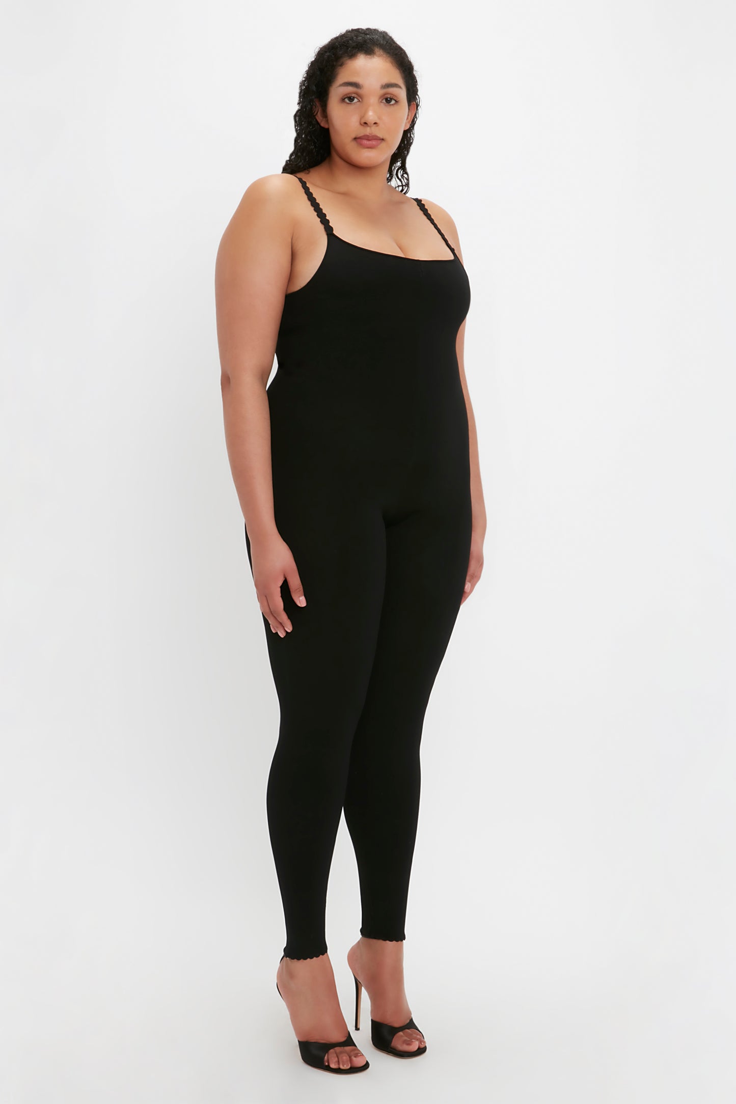 A woman stands against a plain background, wearing the Victoria Beckham VB Body Catsuit in black and black high-heeled shoes. She has a neutral expression.