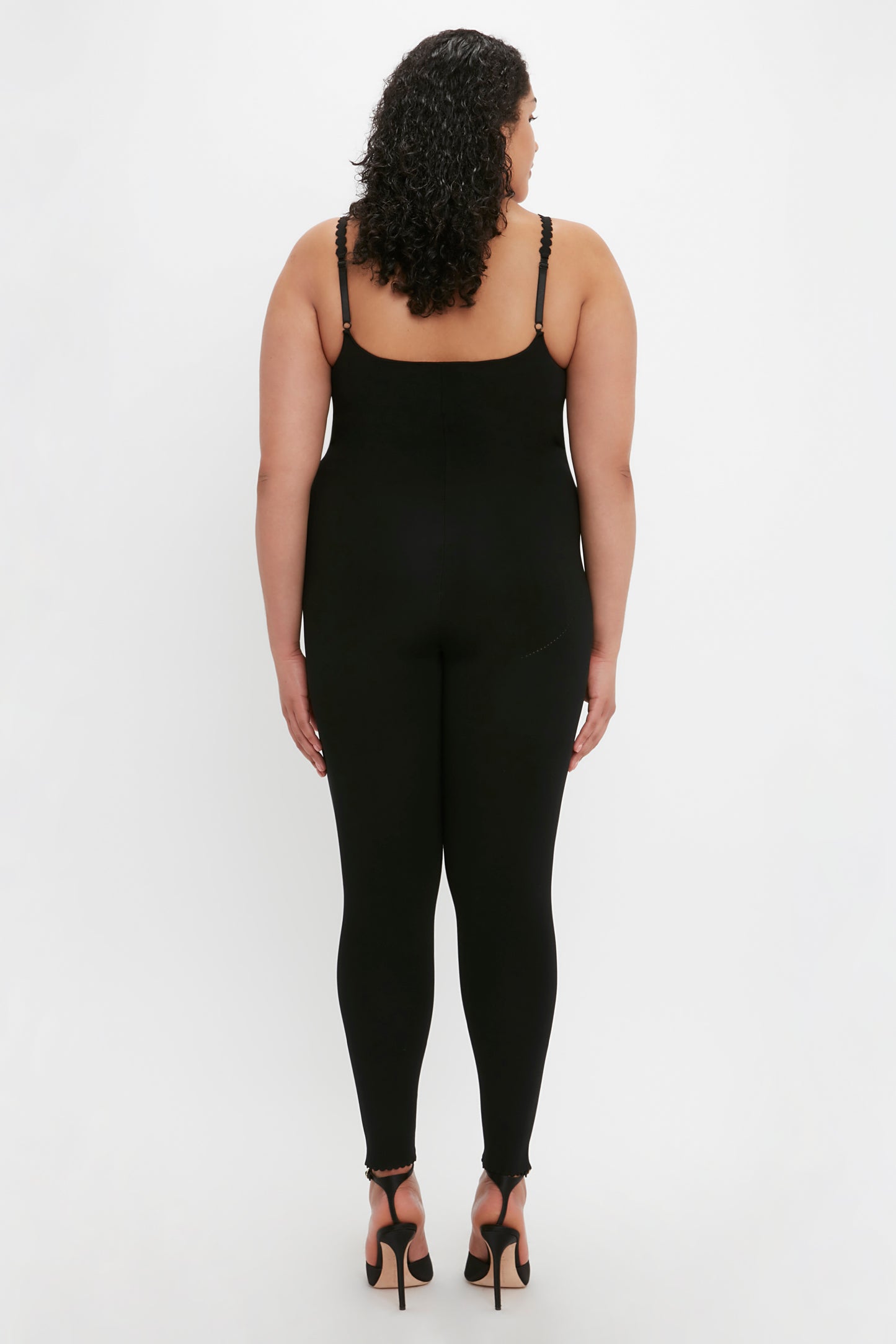 A person with curly hair is standing facing away, wearing the Victoria Beckham VB Body Catsuit in Black and black high-heeled shoes against a plain white background.