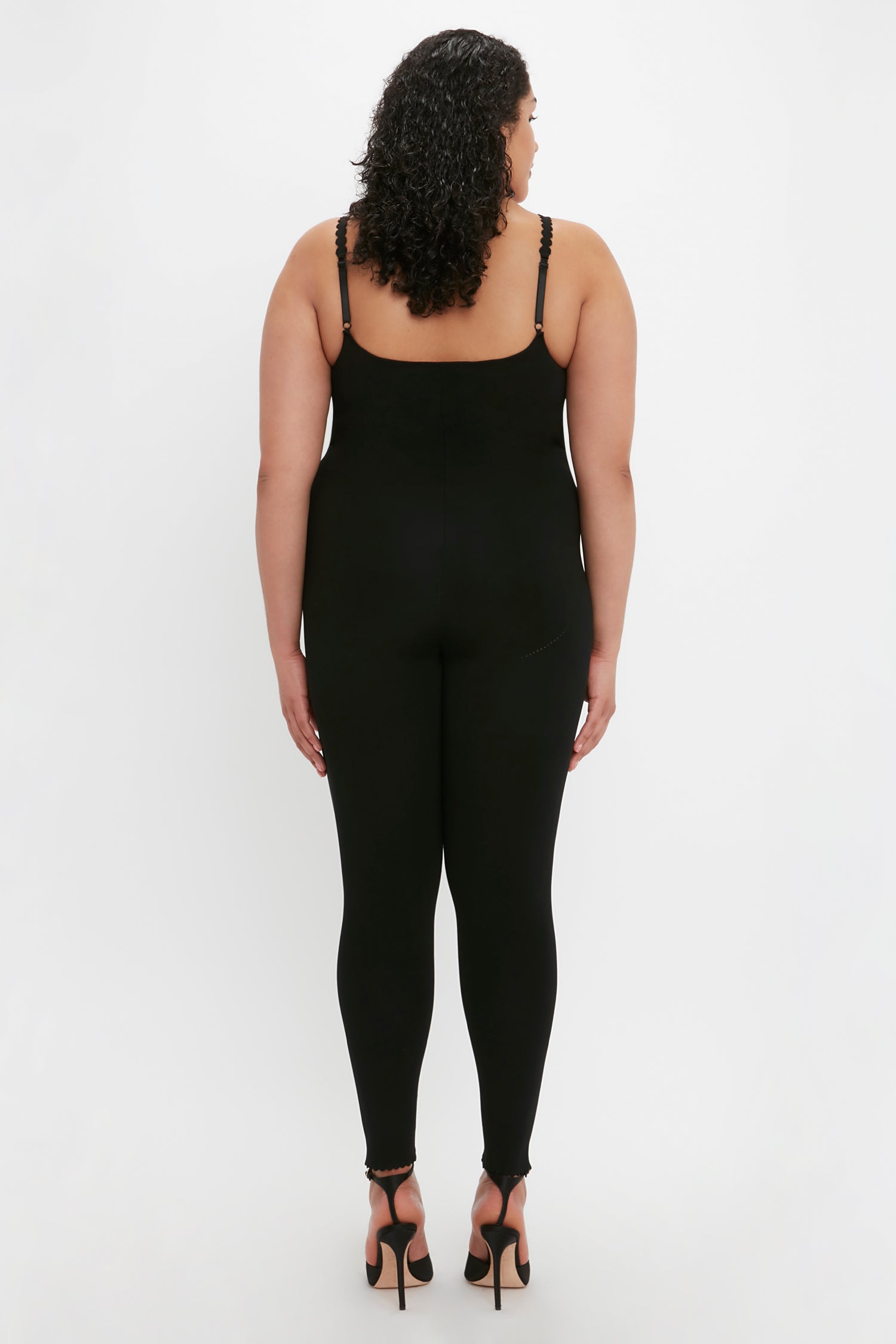 A person with curly hair is standing facing away, wearing the Victoria Beckham VB Body Catsuit in Black and black high-heeled shoes against a plain white background.