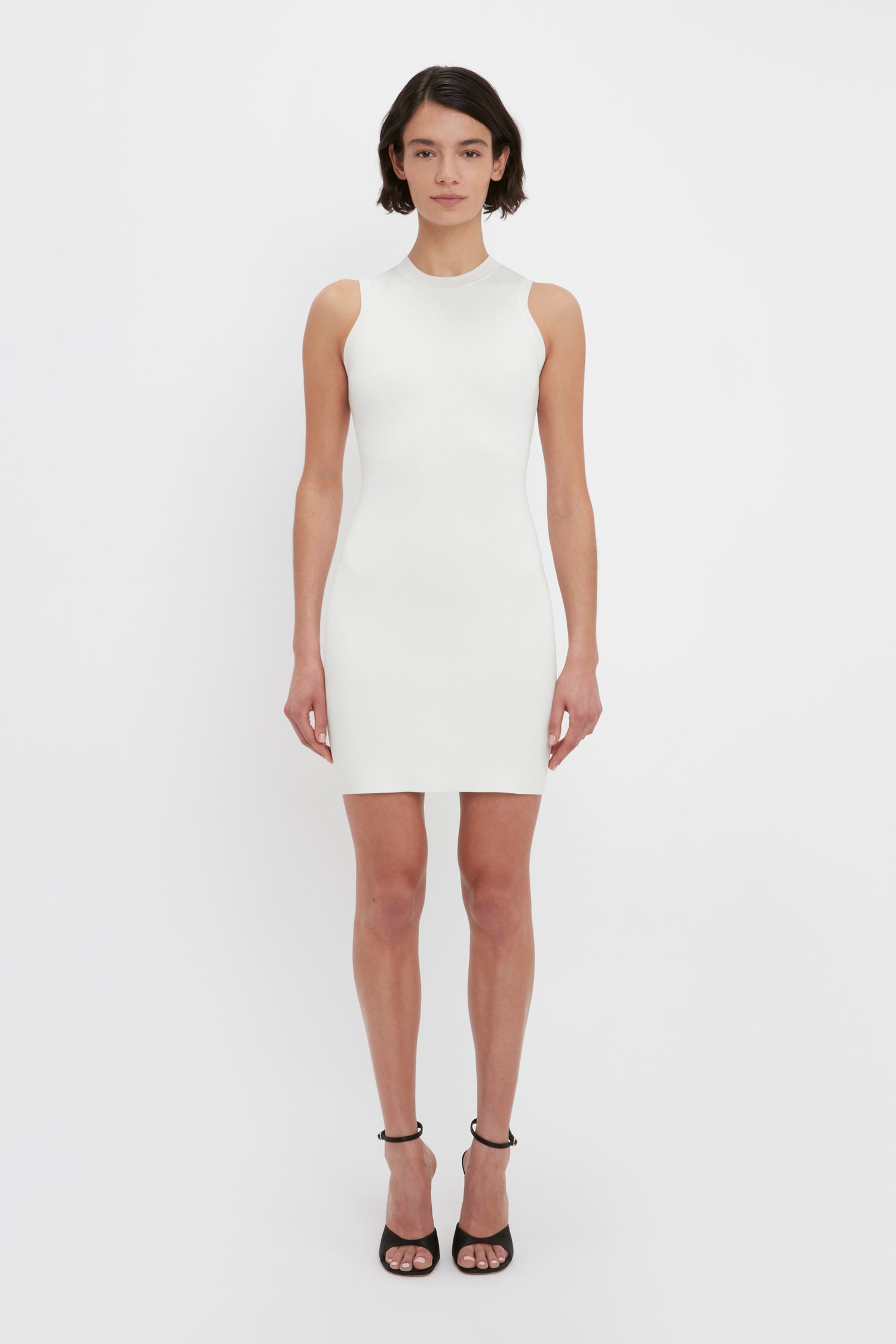 A person stands against a white background wearing the VB Body Mini Dress in White by Victoria Beckham, paired with black high-heeled sandals.